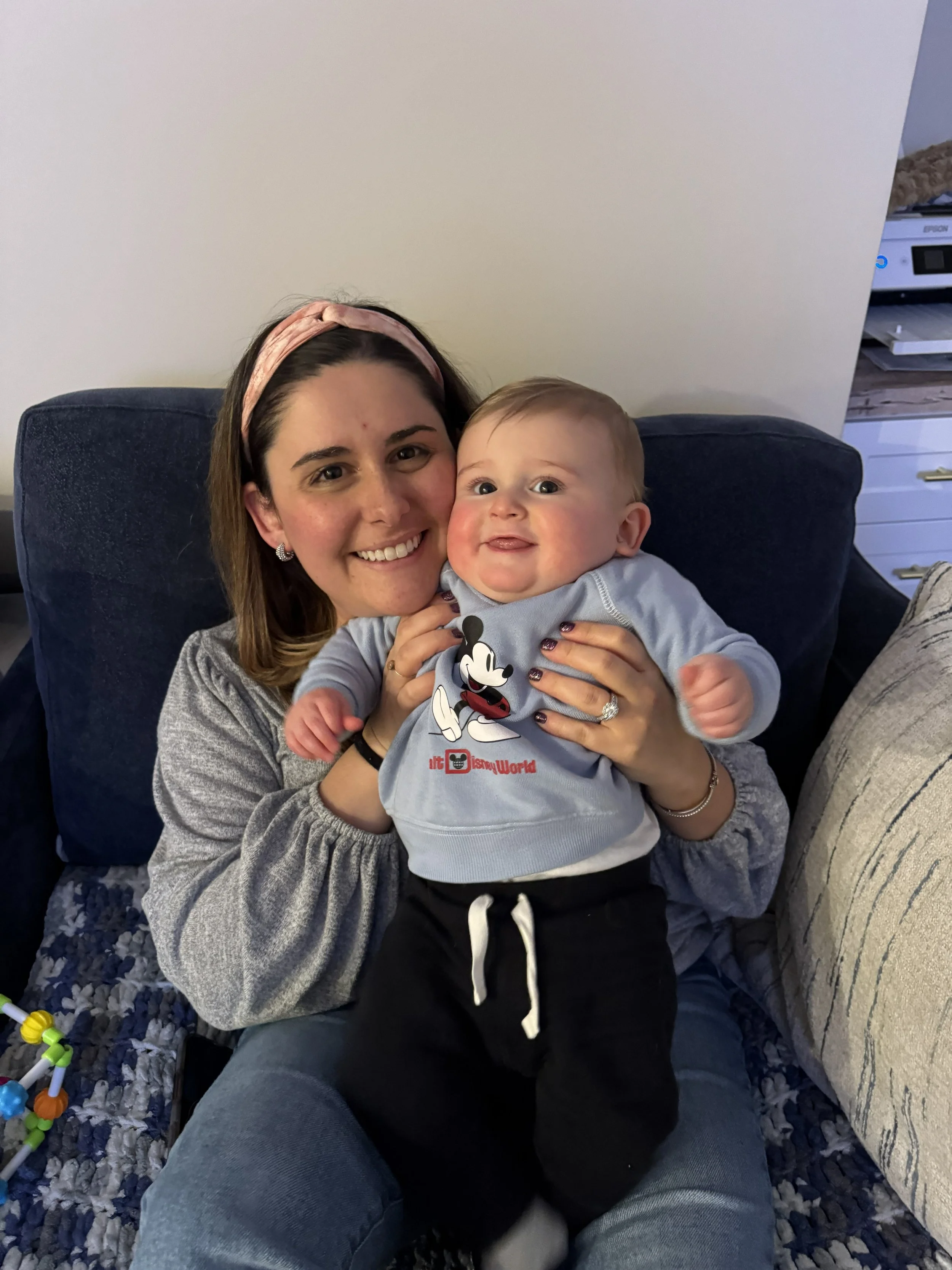A woman with shoulder-length brown hair, wearing a pink headband and a gray top, holding a smiling baby boy with blond hair, blue eyes, and chubby cheeks, dressed in a Mickey Mouse sweatshirt.
