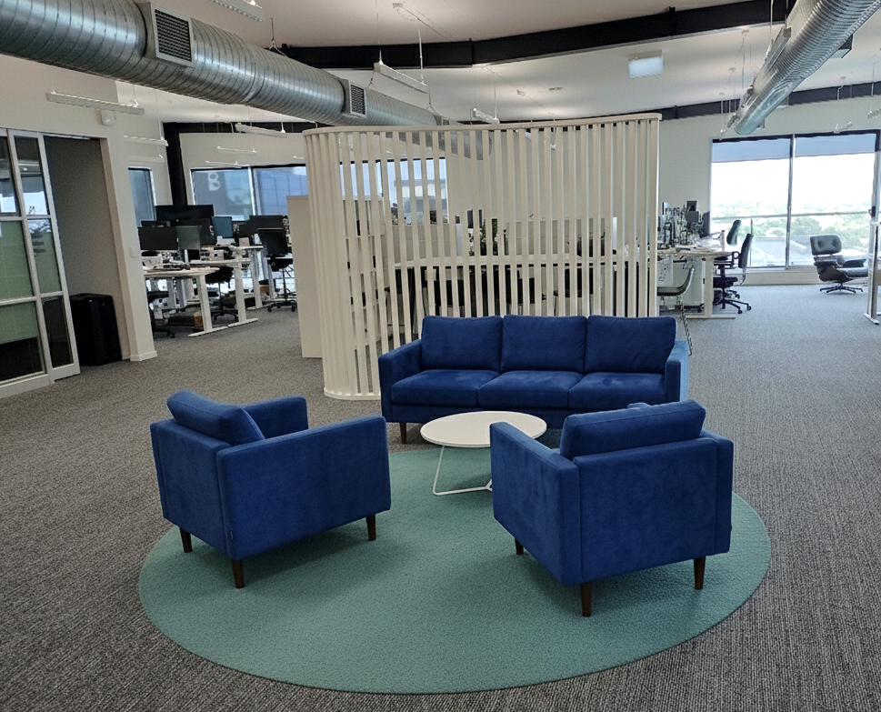 Office lounge area with a blue sofa and two armchairs surrounding a white round coffee table on a green rug, with an open workspace with desks and computers in the background.