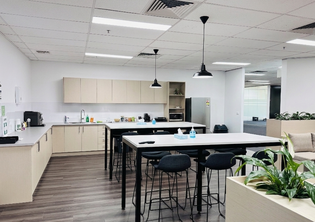 An office communal kitchen area with a coffee maker, microwave, and sink, along with tables and chairs, hand sanitizer bottles, and a sofa with a plant in the foreground.