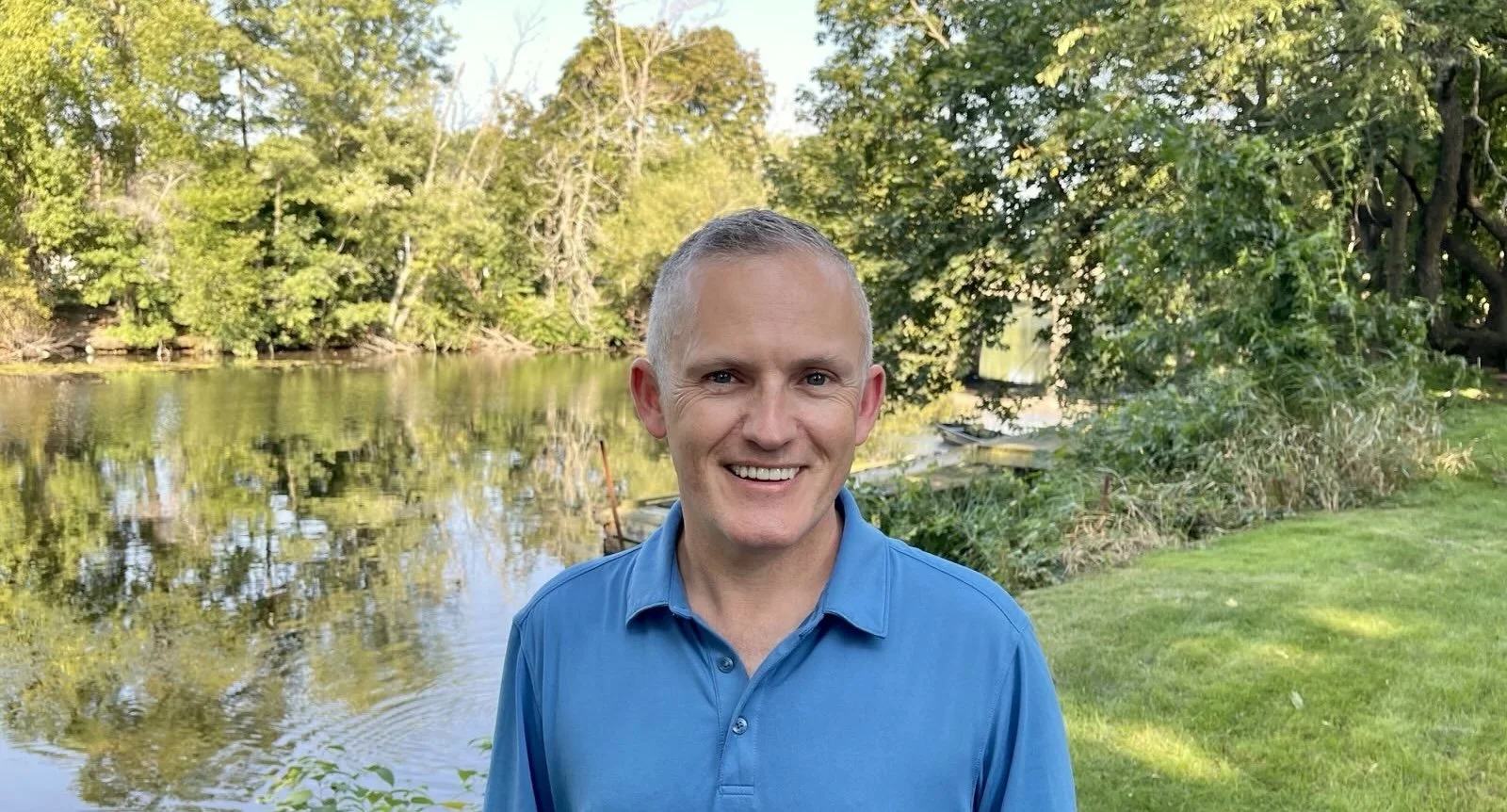 Paul Ruseau, wearing a blue polo shirt smiling outdoors near a river with green trees and grass in the background.