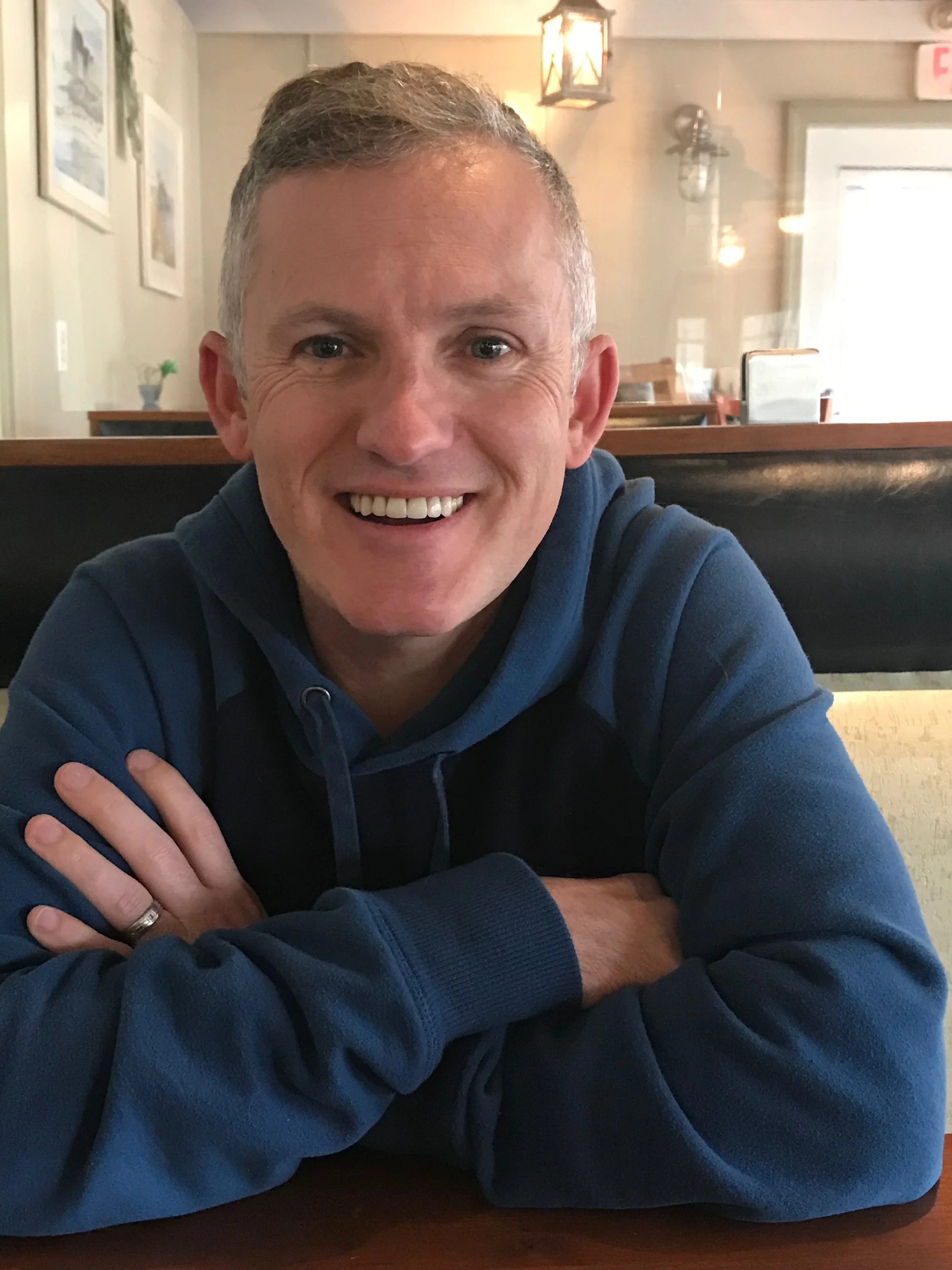 Paul Ruseau, wearing a blue hoodie, sitting at a restaurant table with folded arms, in a cozy indoor setting with wall art and lighting fixtures in the background.