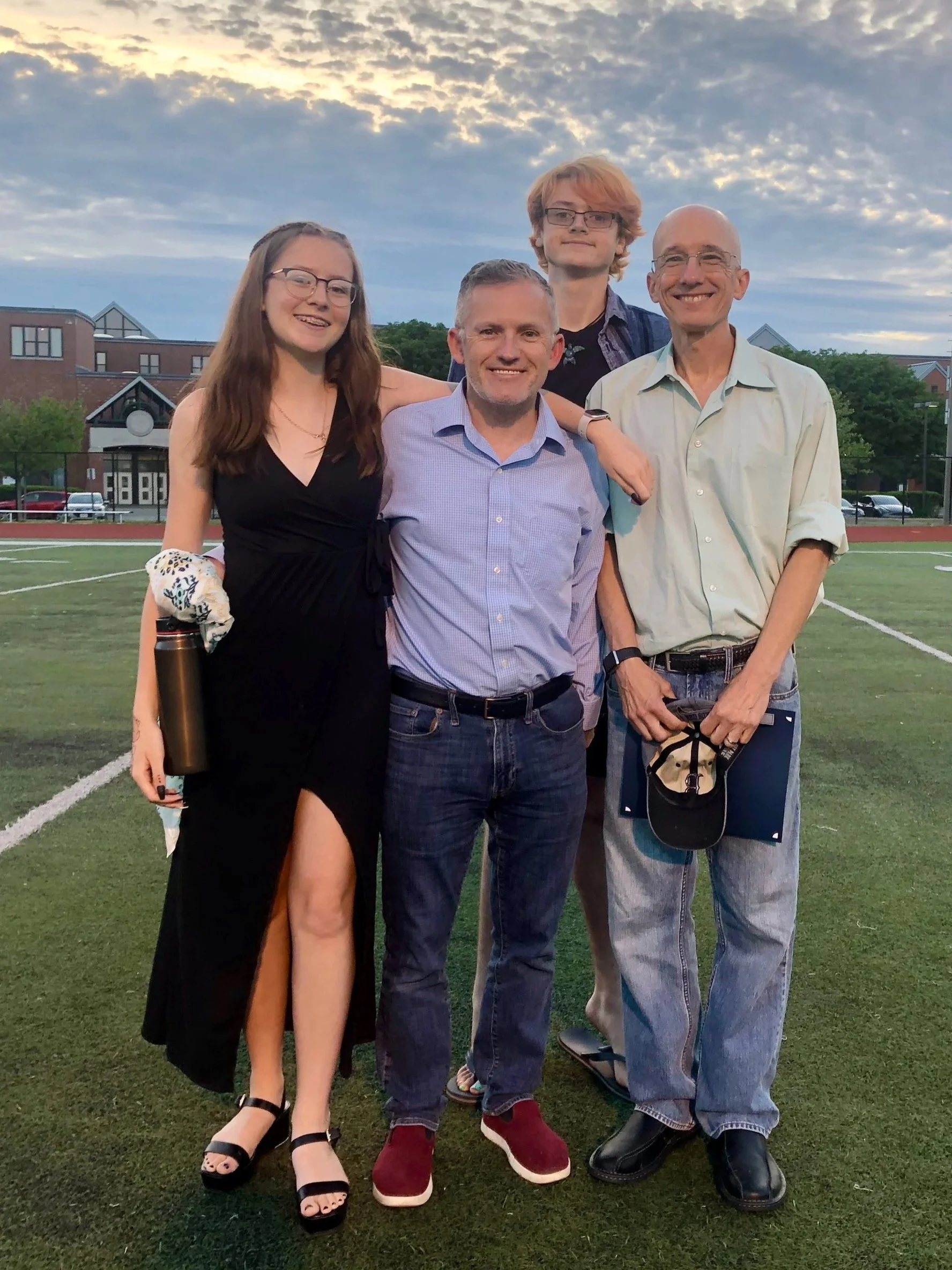 A family of five stands on a football field during sunset, smiling at the camera. The group includes a woman in a black dress with a slit, a man in a blue shirt and jeans, an older man in a light shirt with rolled-up sleeves holding a cap and clipboard, and two young men with glasses, one in a blazer and the other in a dress shirt.
