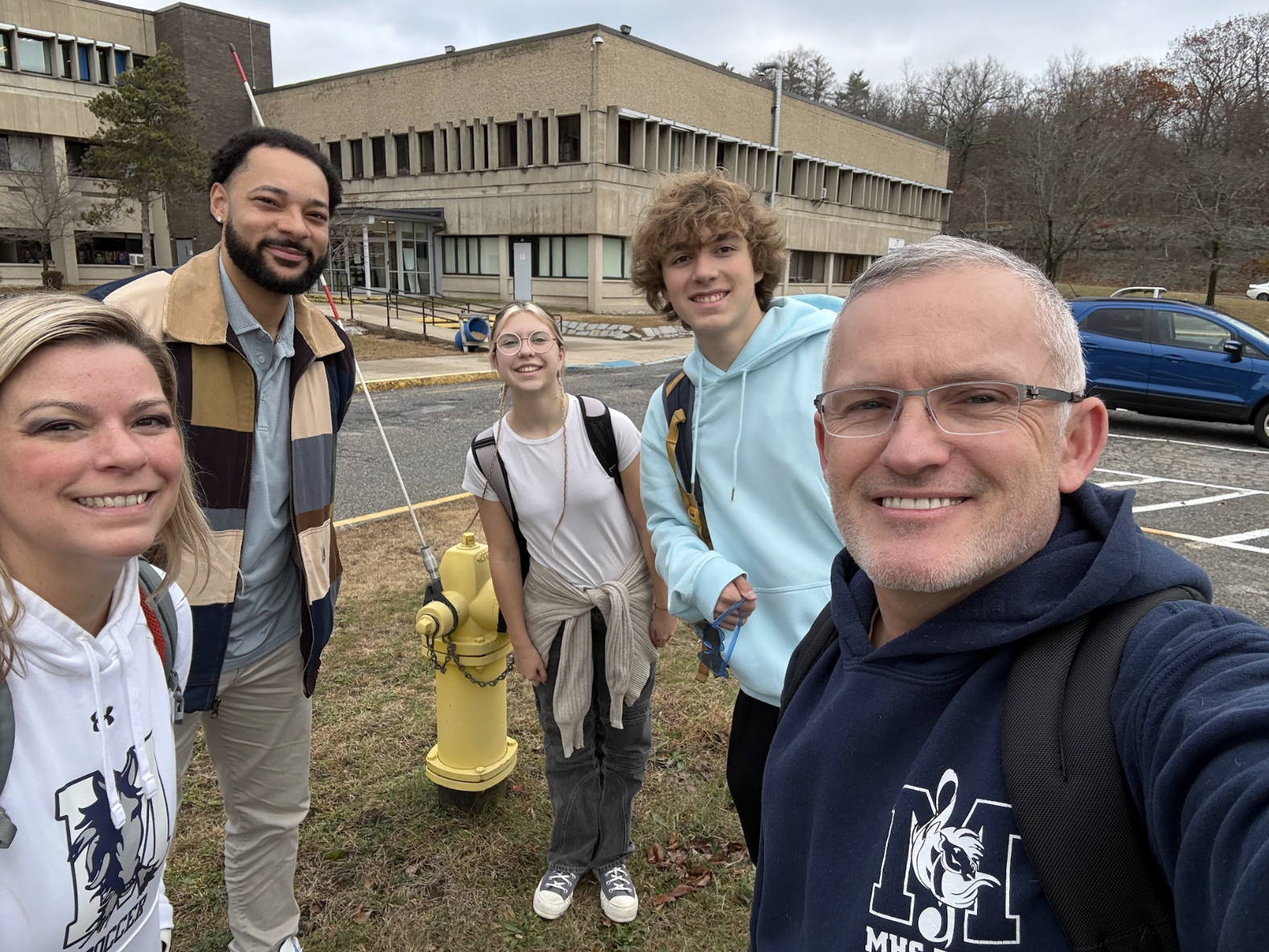 Group of five people smiling outdoors in front of a yellow fire hydrant, with a school building and parked cars in the background.