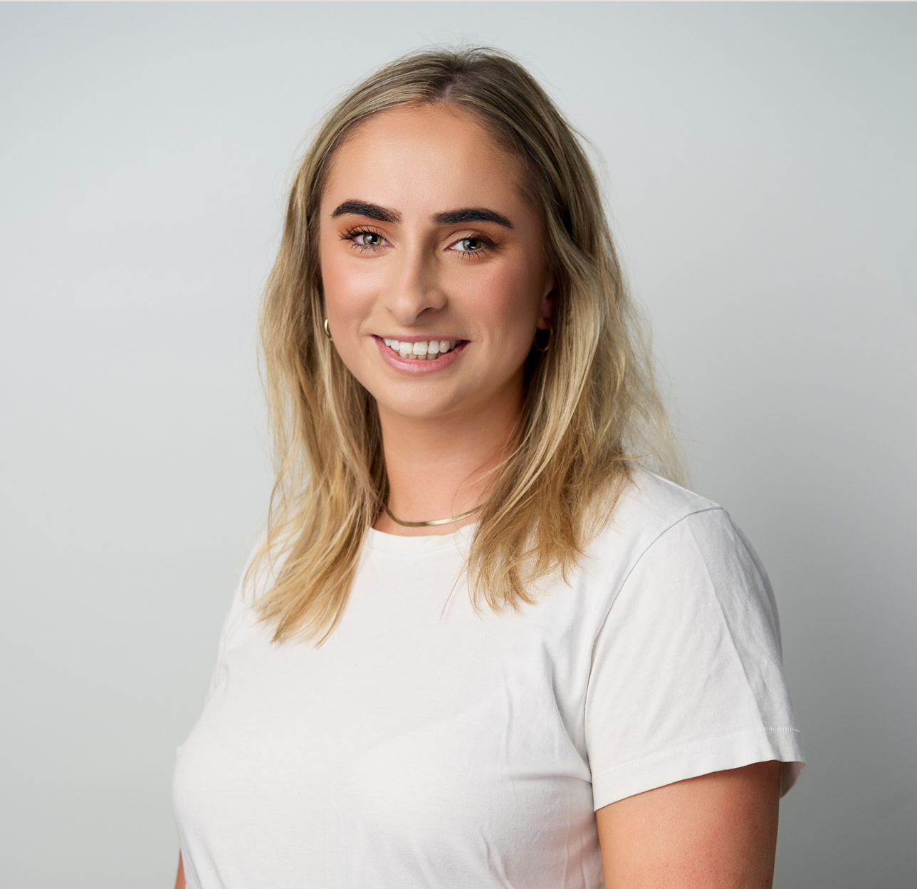 A young woman with light brown hair, light skin, and green eyes smiling, wearing a white t-shirt, gold hoop earrings, and a gold necklace against a light gray background.