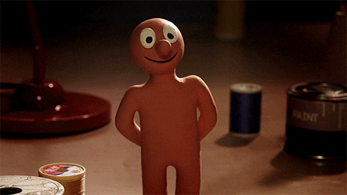 A small humanoid figure made of modeling clay with a round head, large eyes, and a big nose, standing on a surface with various objects around it.