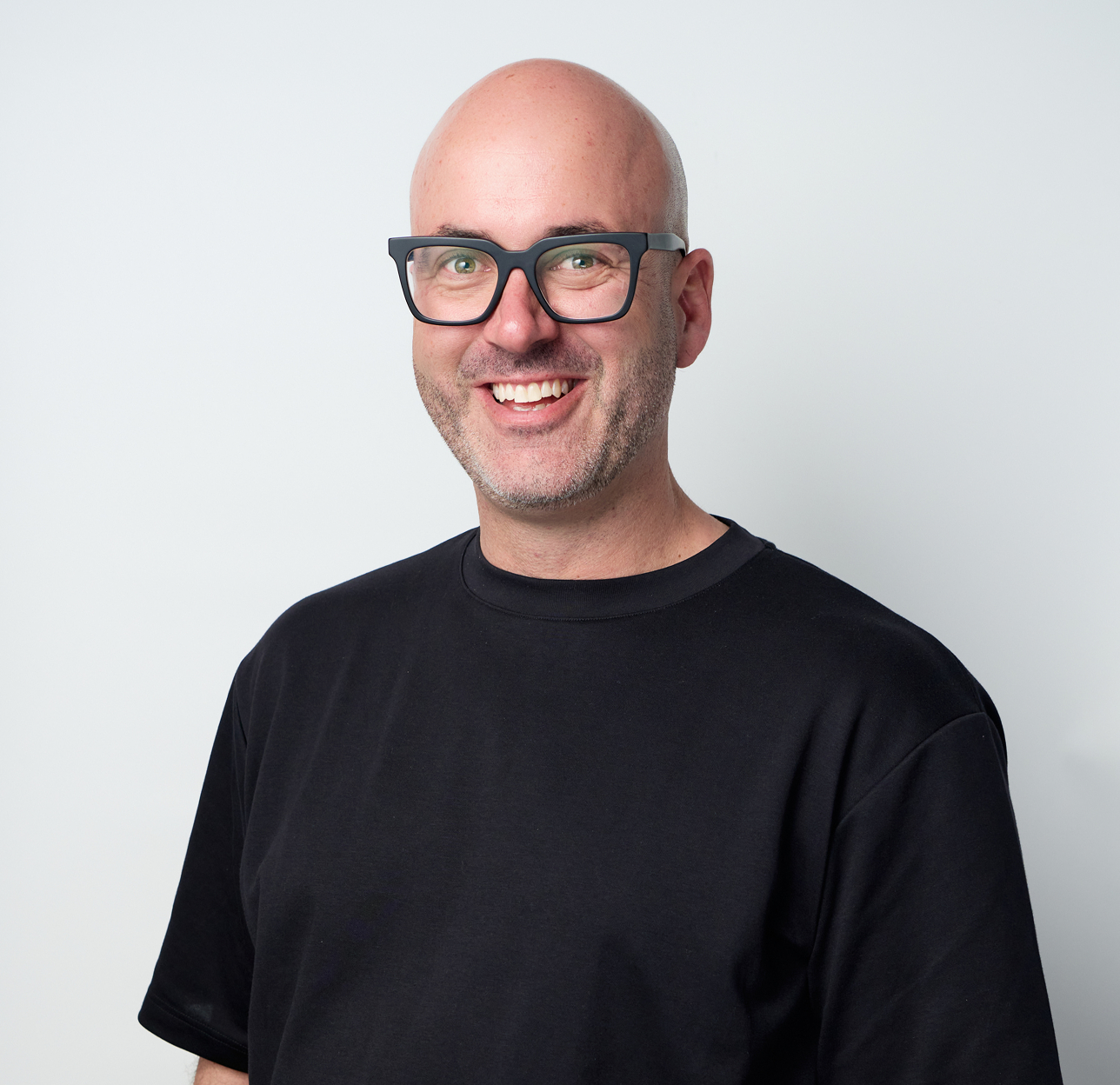 A smiling, bald man with glasses wearing a black T-shirt standing against a plain white background.