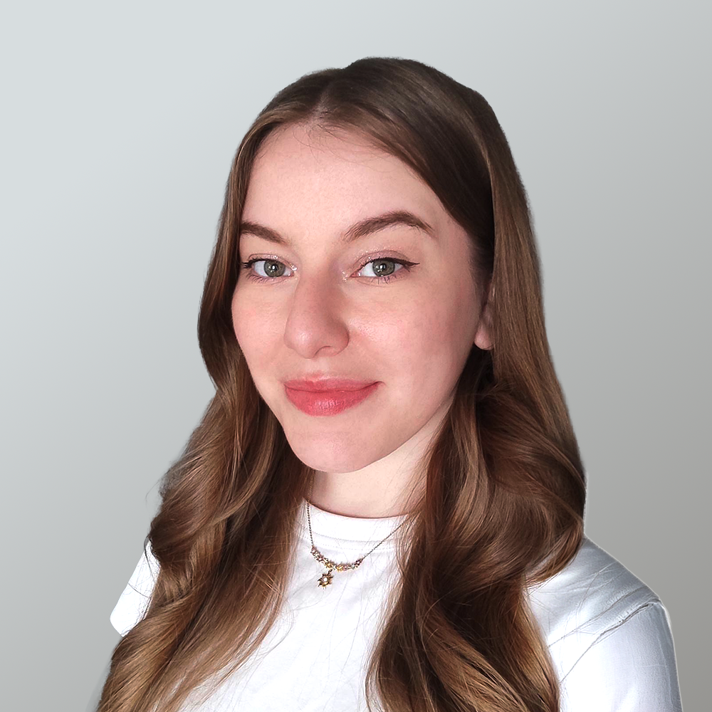 A young woman with long brown hair, light skin, and green eyes, smiling slightly, wearing a white top and layered necklaces, against a plain gray background.
