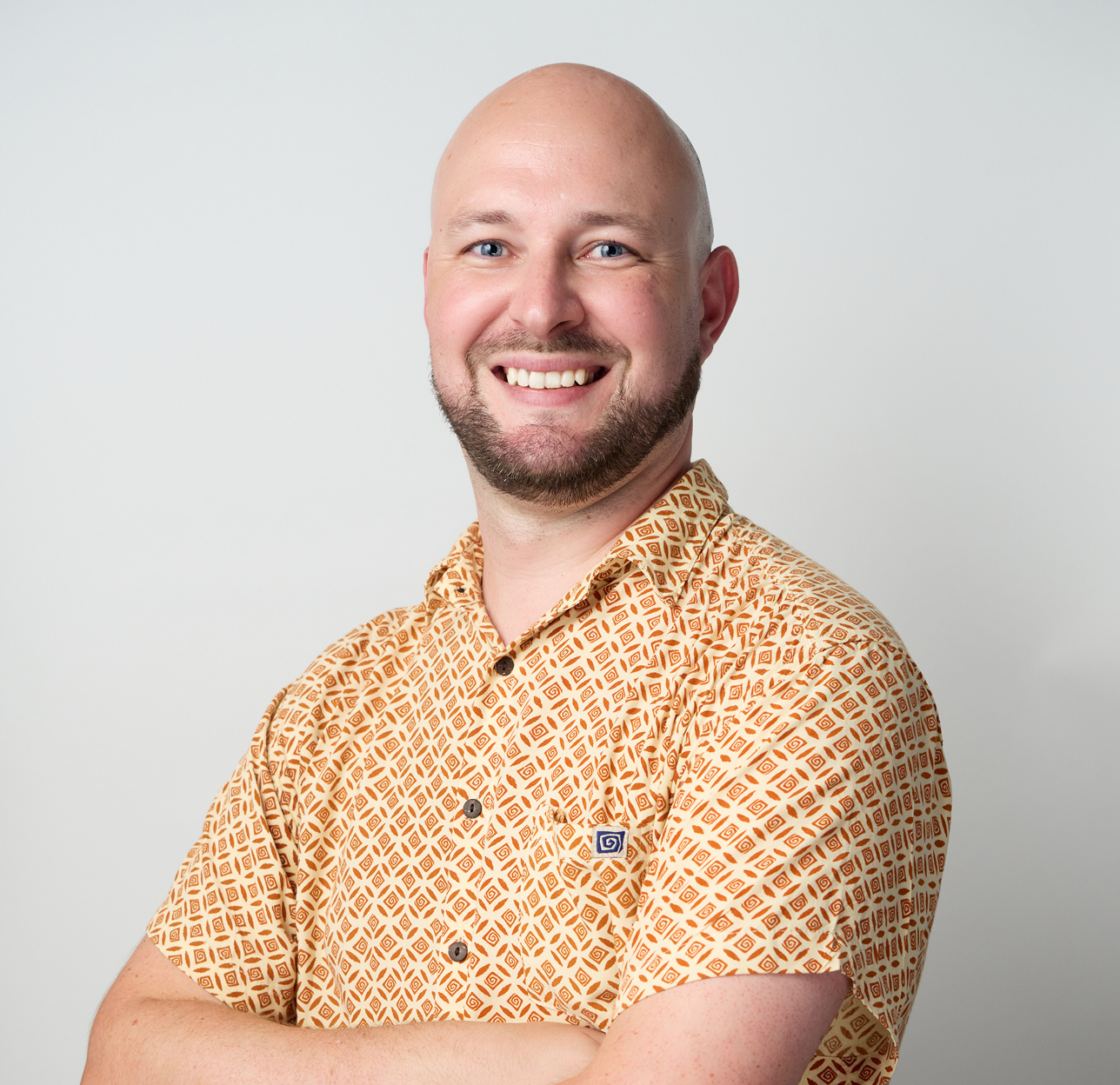 A smiling man with a beard and shaved head wearing an orange patterned short-sleeve shirt, standing against a plain white background.