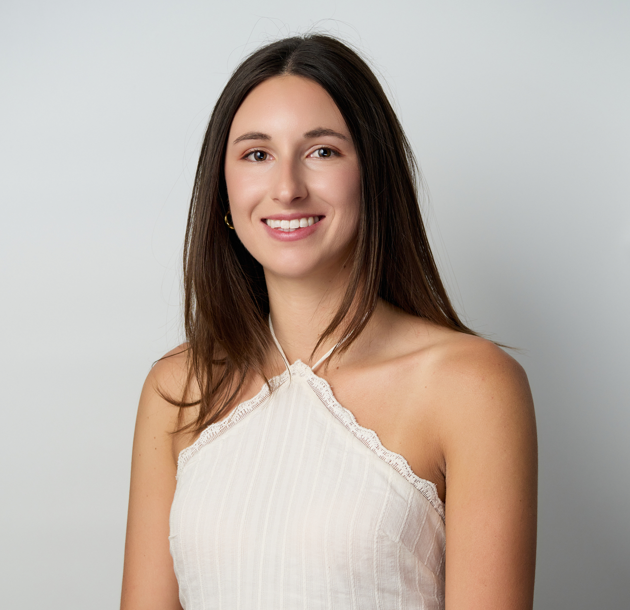 A young woman with long brown hair, smiling, wearing a white sleeveless top, against a plain light gray background.
