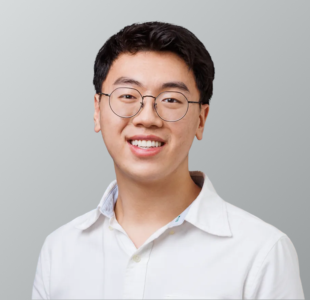 Close-up photo of young man with glasses smiling, wearing a white collared shirt, against a plain gray background.