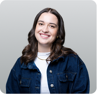 A young woman with shoulder-length dark hair, smiling, wearing a white top and a blue denim jacket against a plain light gray background.