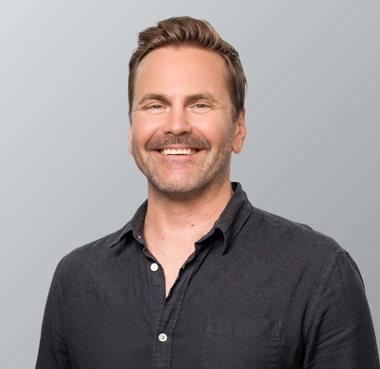 A smiling man with short brown hair, a beard, and wearing a dark button-up shirt, posed against a plain light gray background.