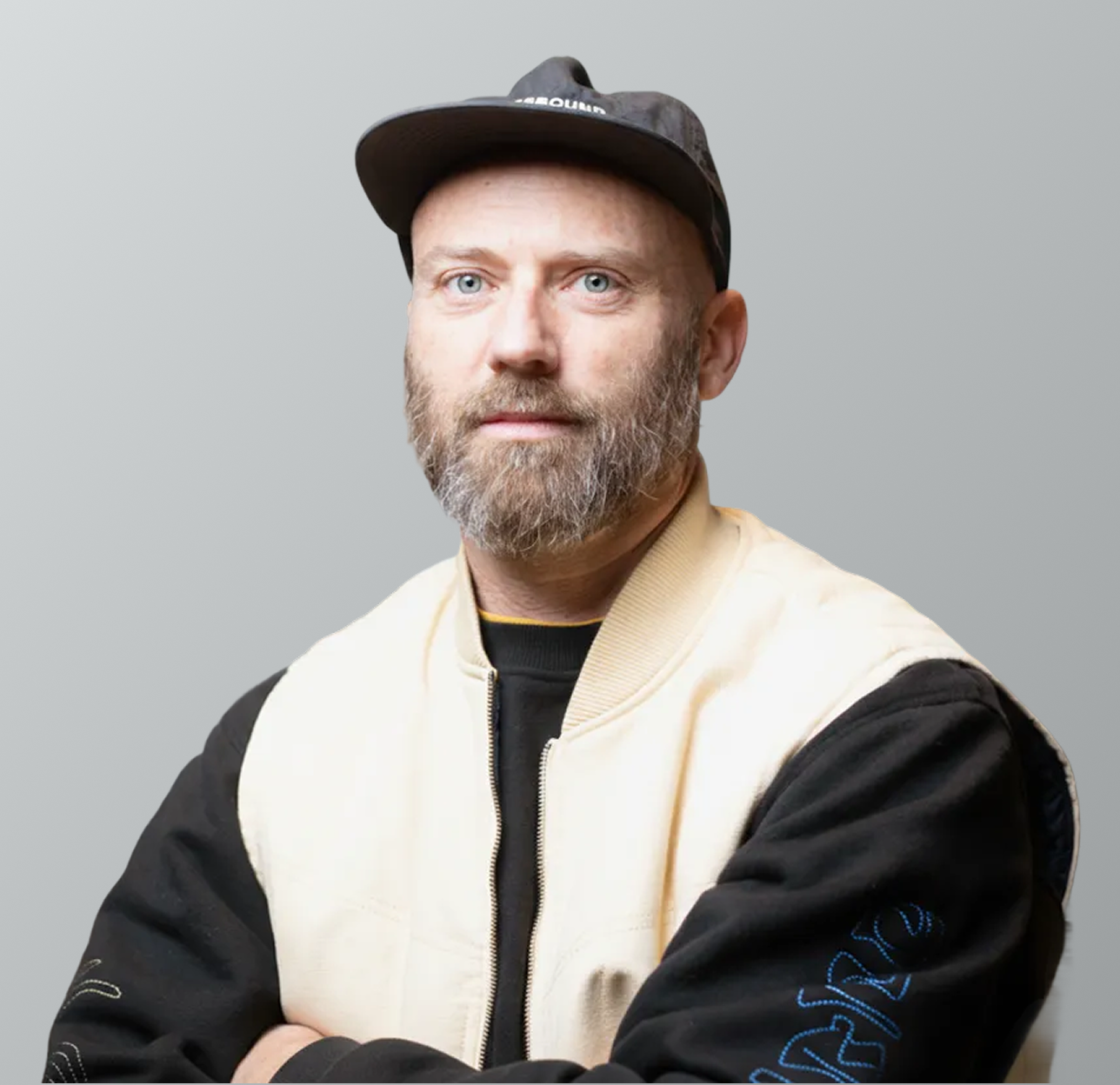 A man with a beard wearing a black cap, a beige jacket with black sleeves, and a black shirt.