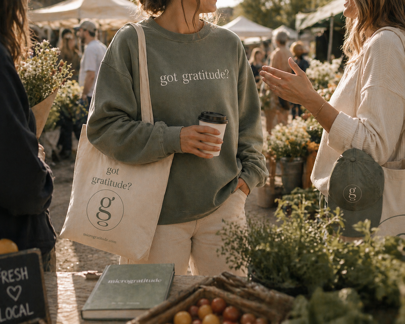 People at an outdoor market engaging in conversation. One person is holding a coffee cup and wearing a gray sweatshirt with the text "got gratitude?". They have a tote bag with the same phrase and a logo reading "g" on it. The market stall displays flowers and vegetables.