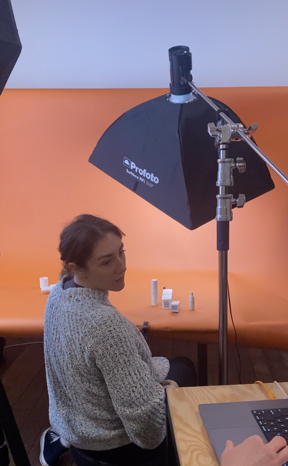 A woman sitting at a table in a photography studio, with various small bottles and boxes in front of her. She is looking over her shoulder, with studio lighting equipment overhead and an orange backdrop behind her.