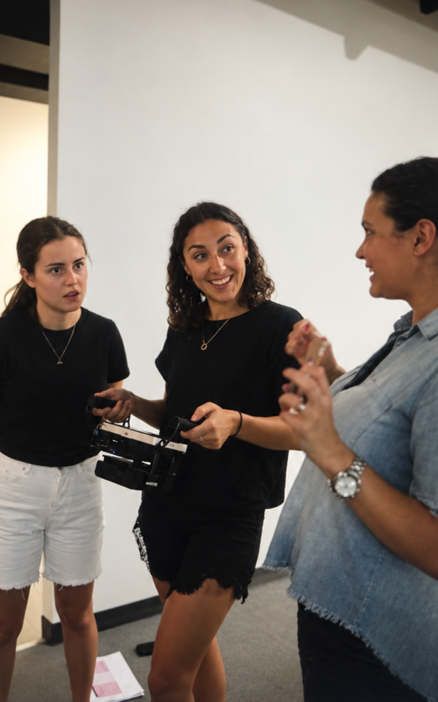 Three women engaged in conversation in an indoor setting, with one woman smiling and holding a camera.