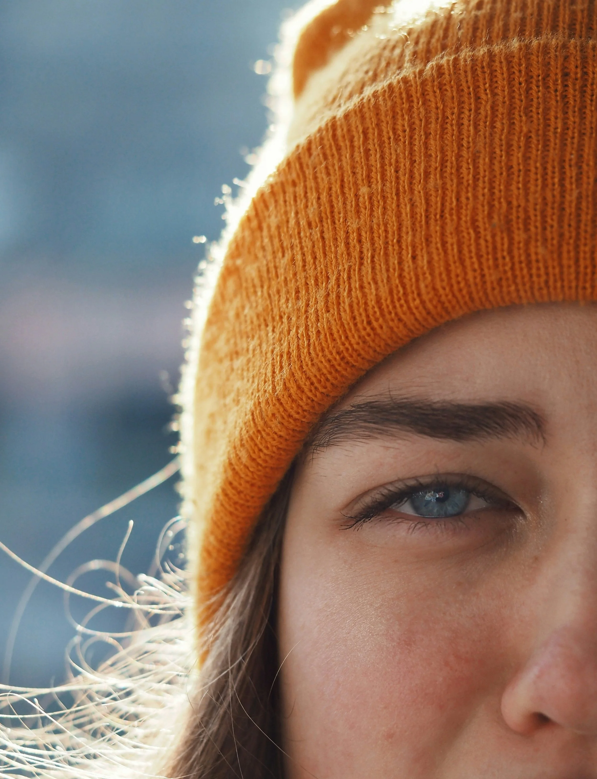 Close-up of a person wearing an orange knit beanie, focusing on their eye and part of their face, with sunlight highlighting the person's hair and the beanie.