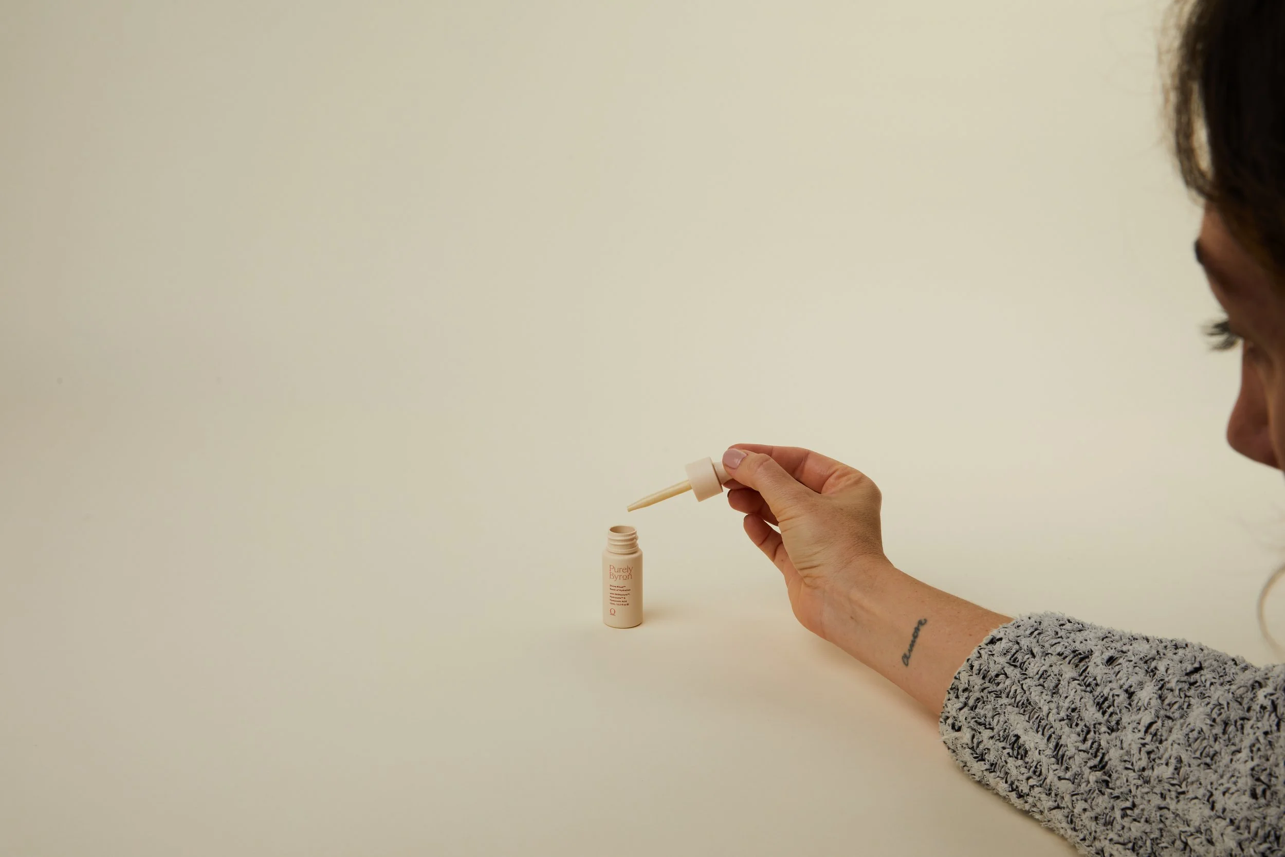 A woman with a tattooed arm is holding a dropper above a bottle of skincare product labeled Purely Byron, against a plain light beige background.