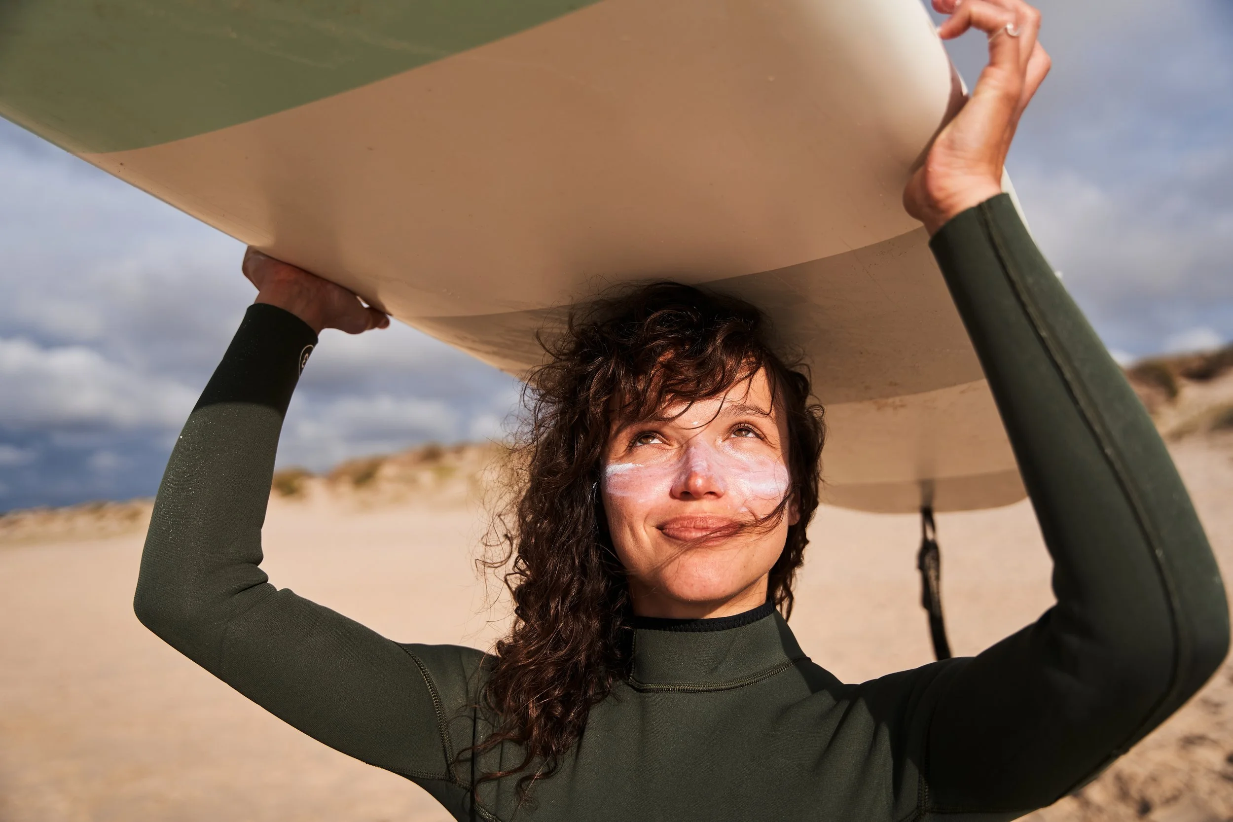 A surfer holding a surfboard on her shoulders on a sandy beach, with cloudy sky in the background.
