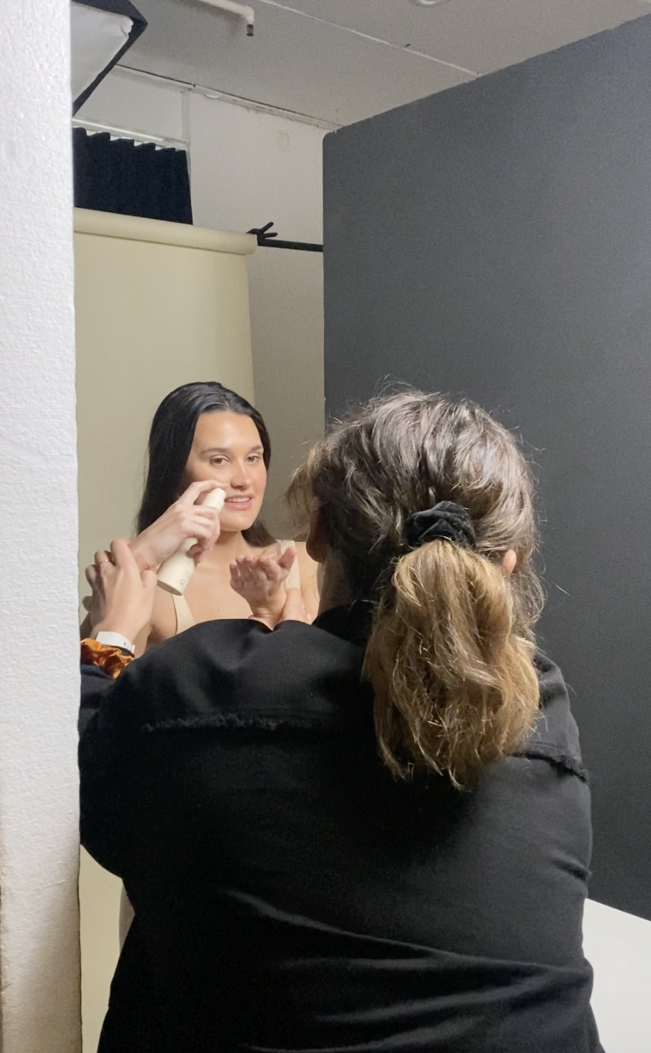 A woman is applying makeup or skincare to her face in front of a mirror while another woman assists her in a studio or dressing room.