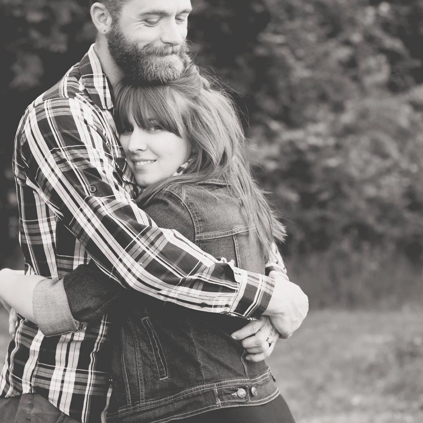 A black and white photo of a happy couple, Chris Cole and Kristina Cole, hugging outdoors, with trees in the background.