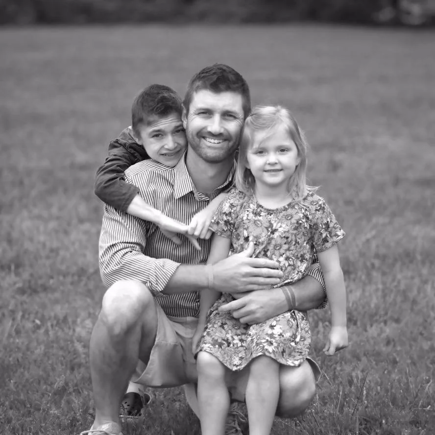 Black and white photo of a man, Josh Helmley, with two children outdoors on grass, all smiling, with the man kneeling and embracing the girl sitting on his knee while the boy is on the man's shoulder.