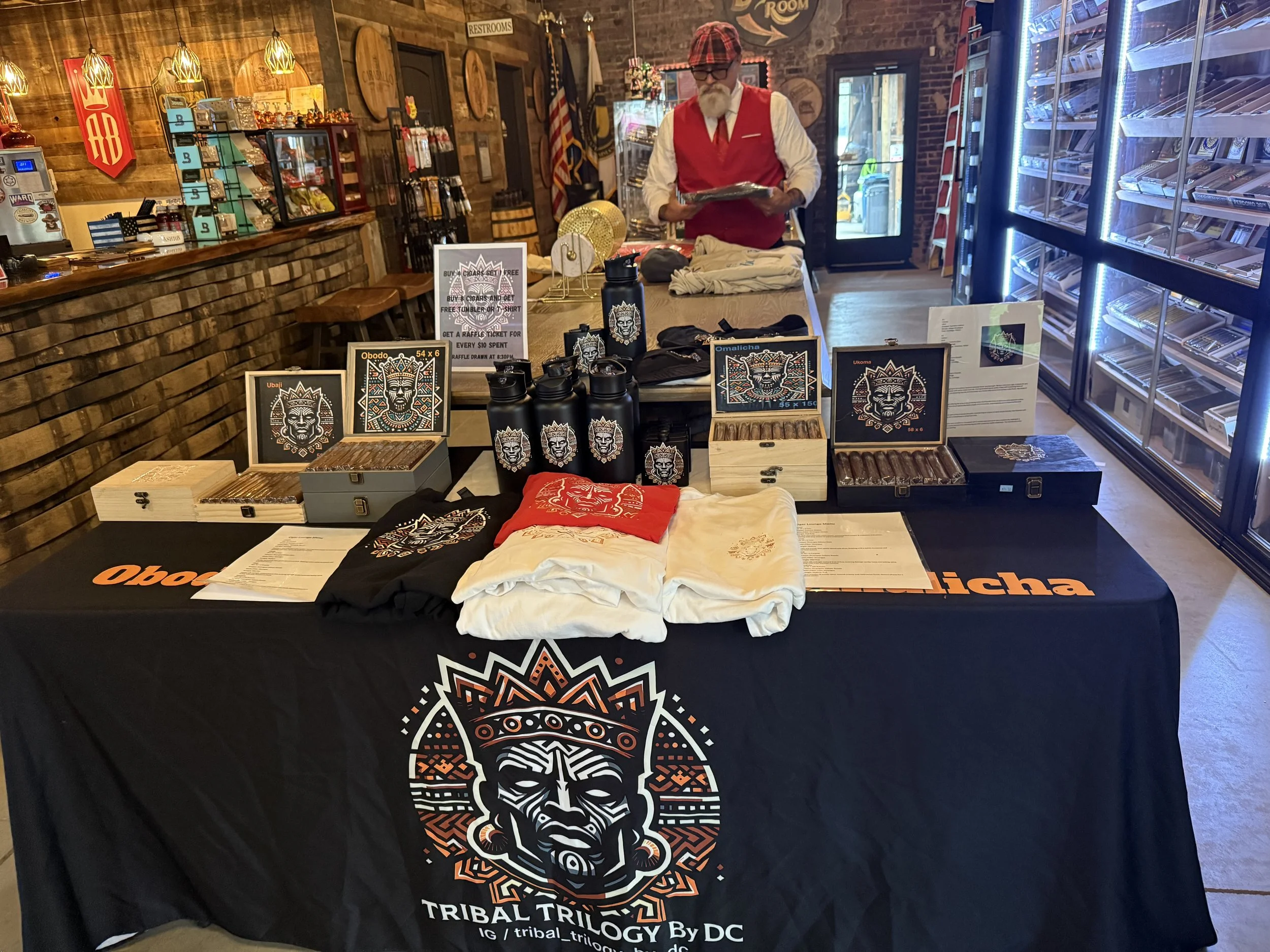 A table covered with tribal-themed merchandise, including T-shirts, posters, water bottles, and cigars, set up inside a store with brick walls, wooden decor, and a man in a red vest and cap standing behind the table.