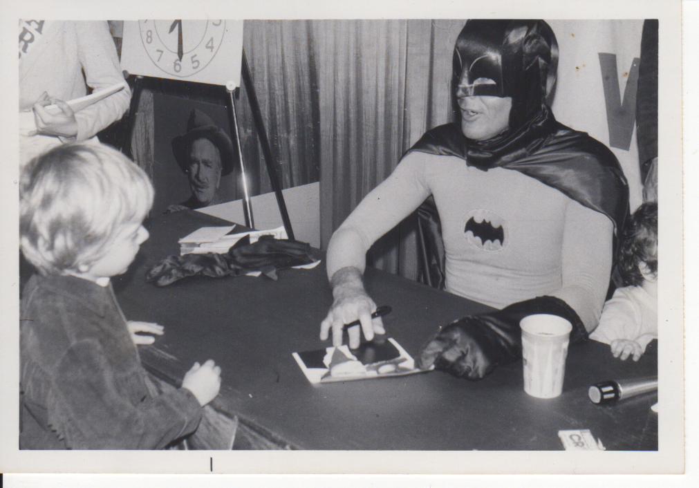 A person dressed as Batman sitting at a table with children, engaging in a friendly interaction at a convention or event. Batman costume includes a mask, cape, and chest logo.