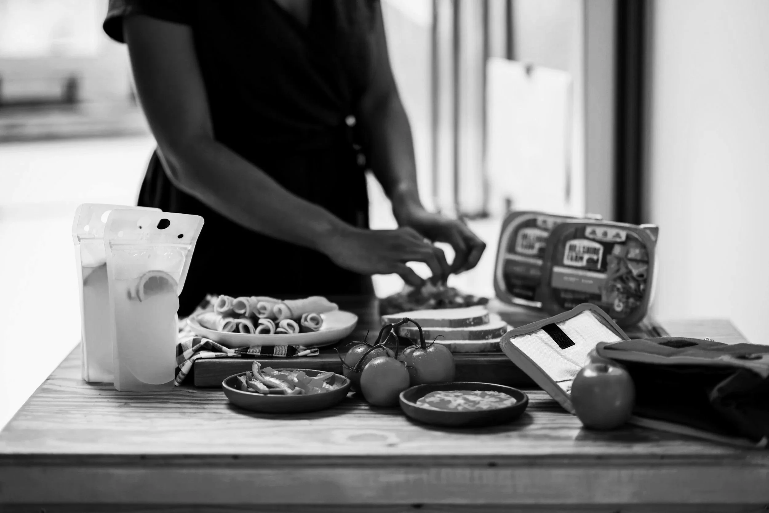 Person preparing food on a wooden table with various plates of sliced vegetables, tomatoes, and a package of food, in a kitchen setting.