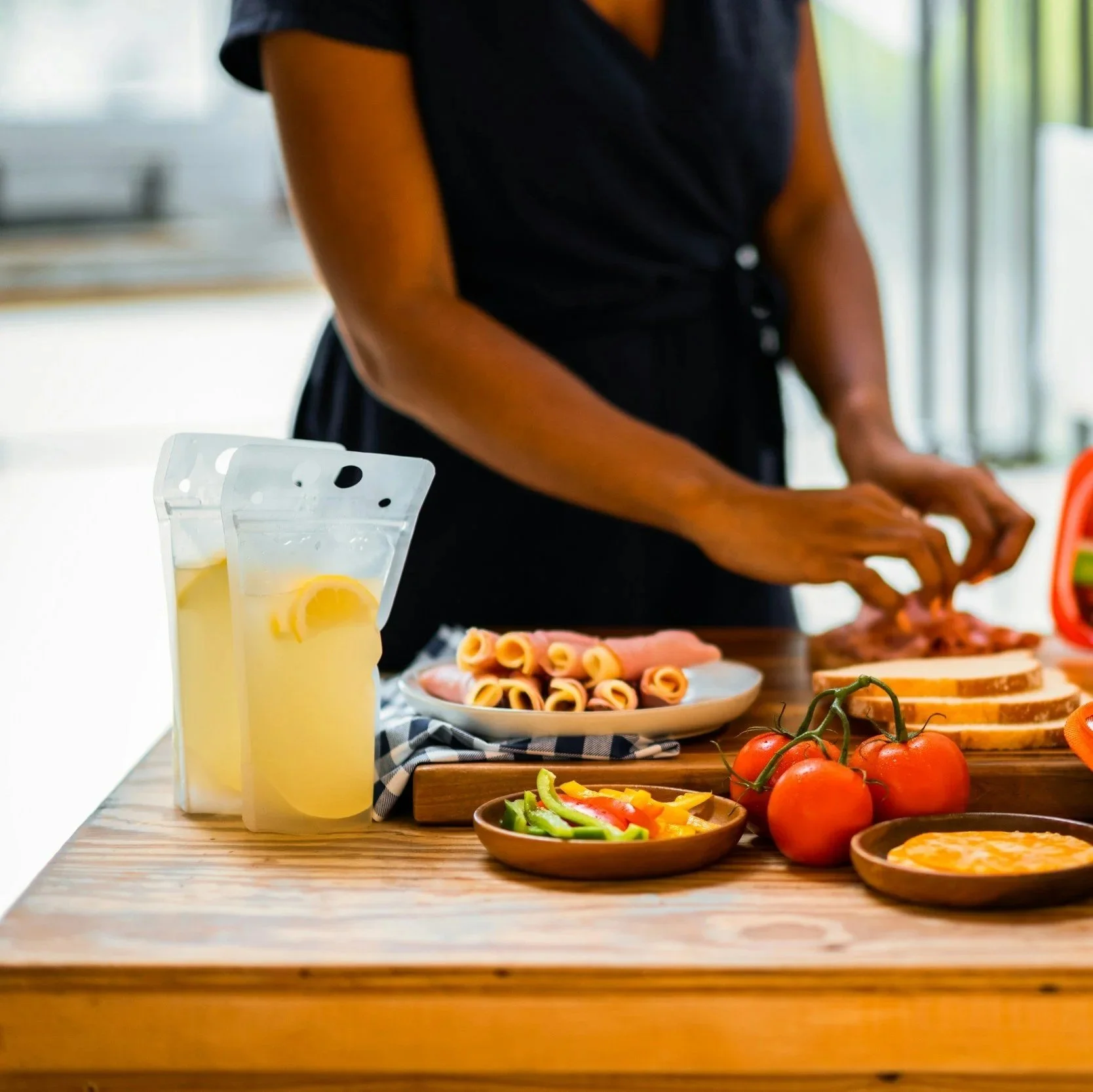 Woman preparing sandwiches on a wooden kitchen table with fresh tomatoes, sliced vegetables, and drinks nearby.