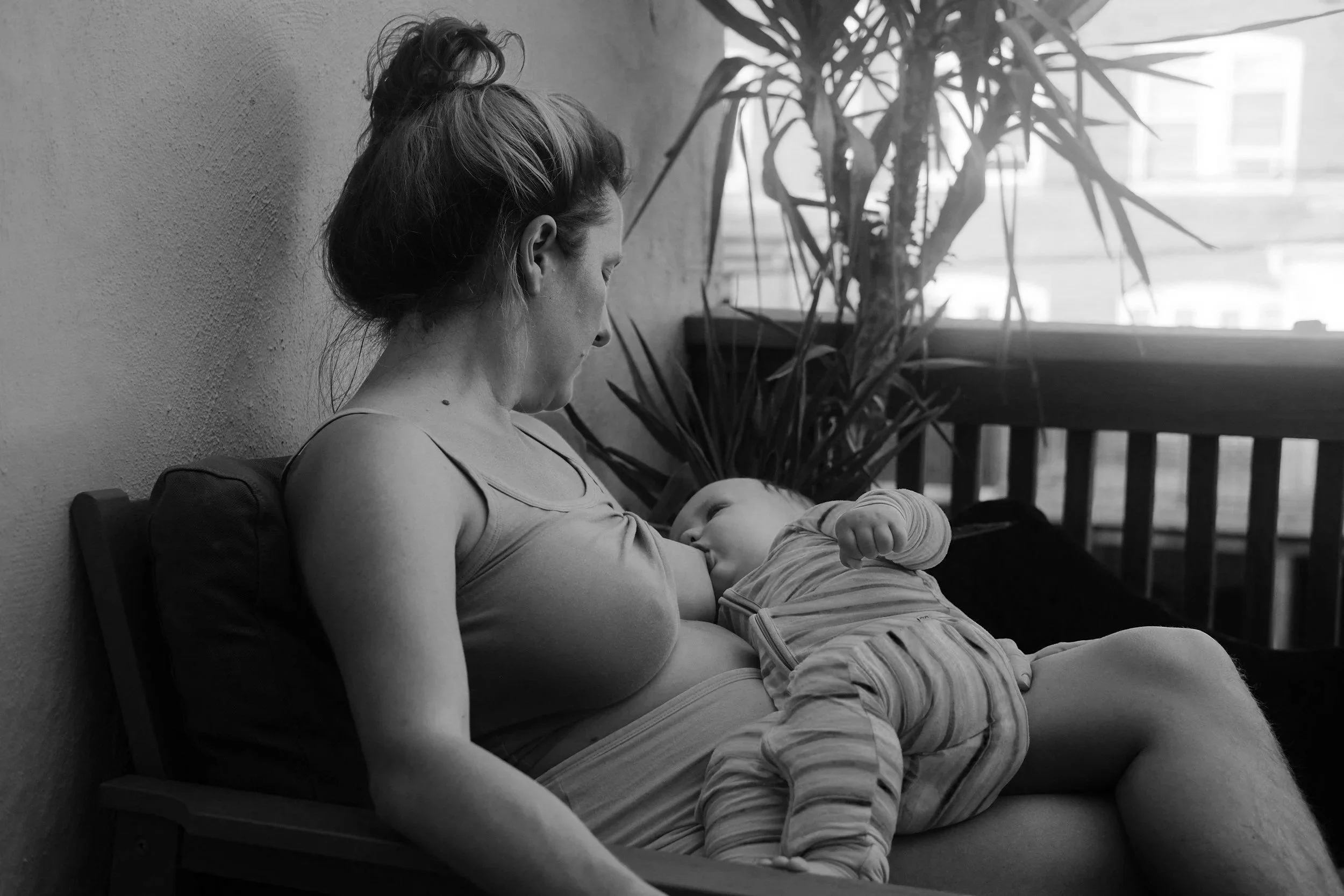 A woman is sitting on a bench, breastfeeding a baby. There is a window and a plant in the background. The image is in black and white.