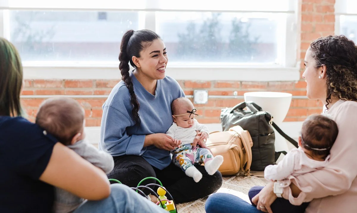 Group of women and babies sitting on the floor in an indoor space with large windows behind them, engaging in conversation.