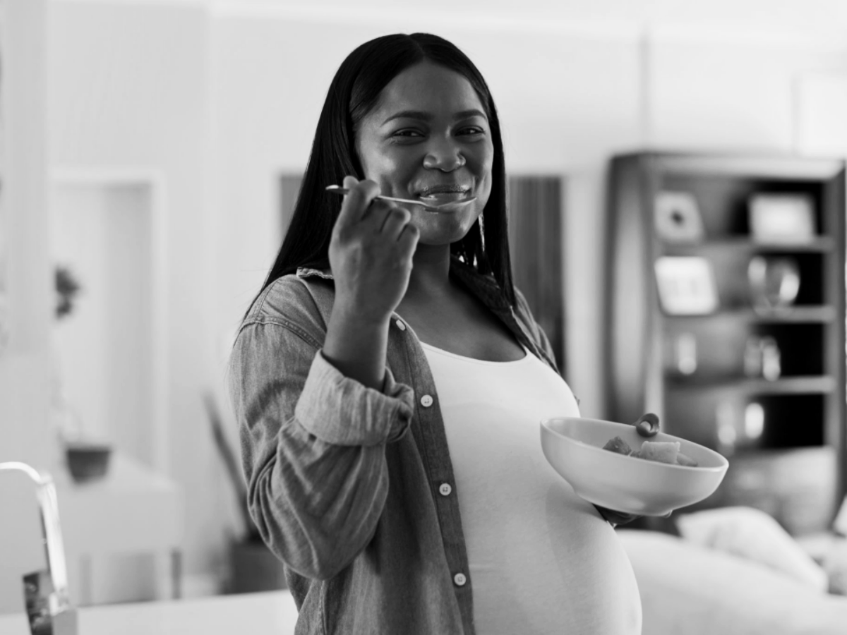 A pregnant woman smiling while holding a bowl of fruit and a spoon in a room, in black and white.