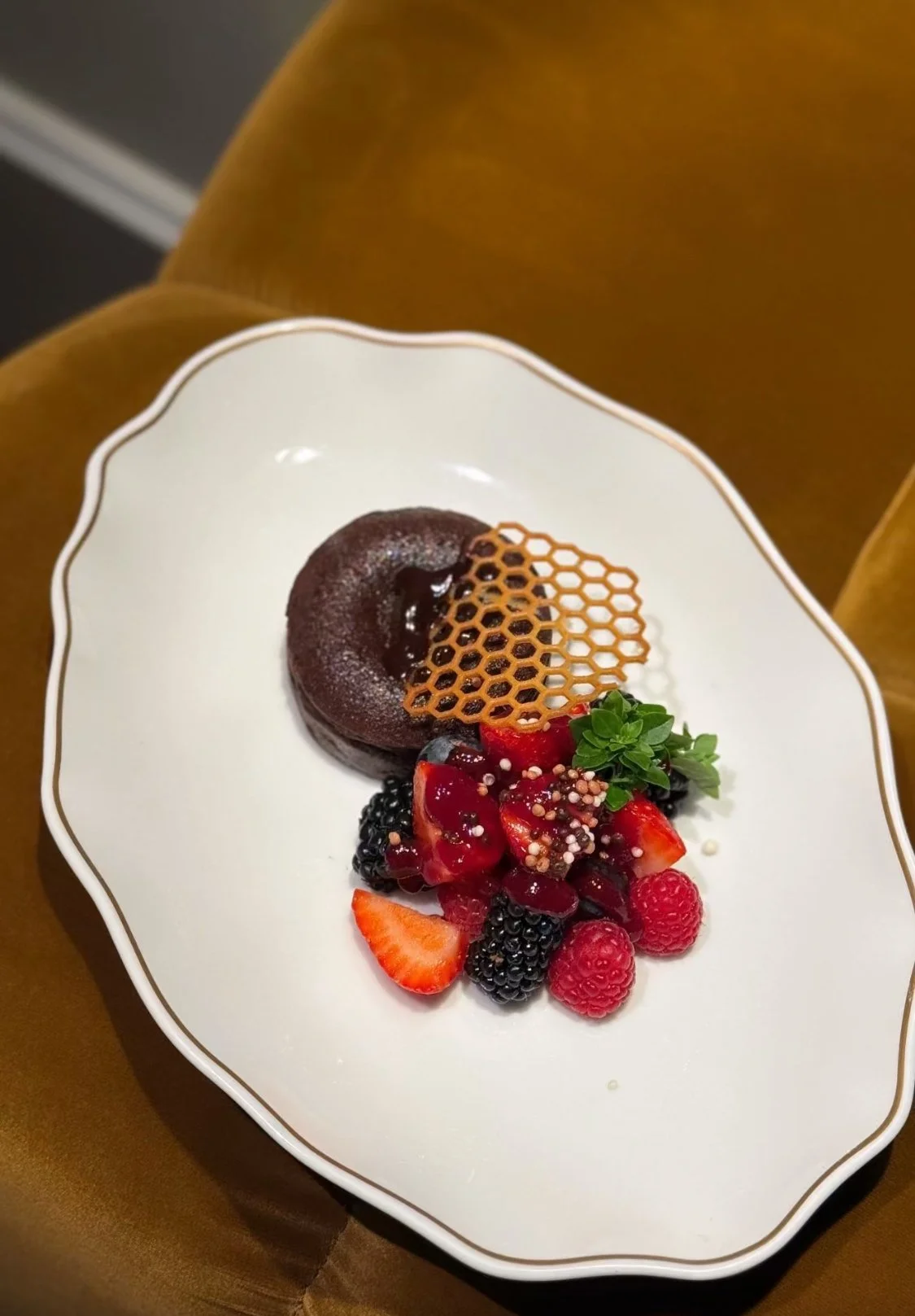 Assiette de dessert avec un gâteau au chocolat, un caramel en forme de nid d'abeille, des fruits rouges, une branche de menthe et des perles de sucre, sur une assiette blanche avec un rebord doré.
