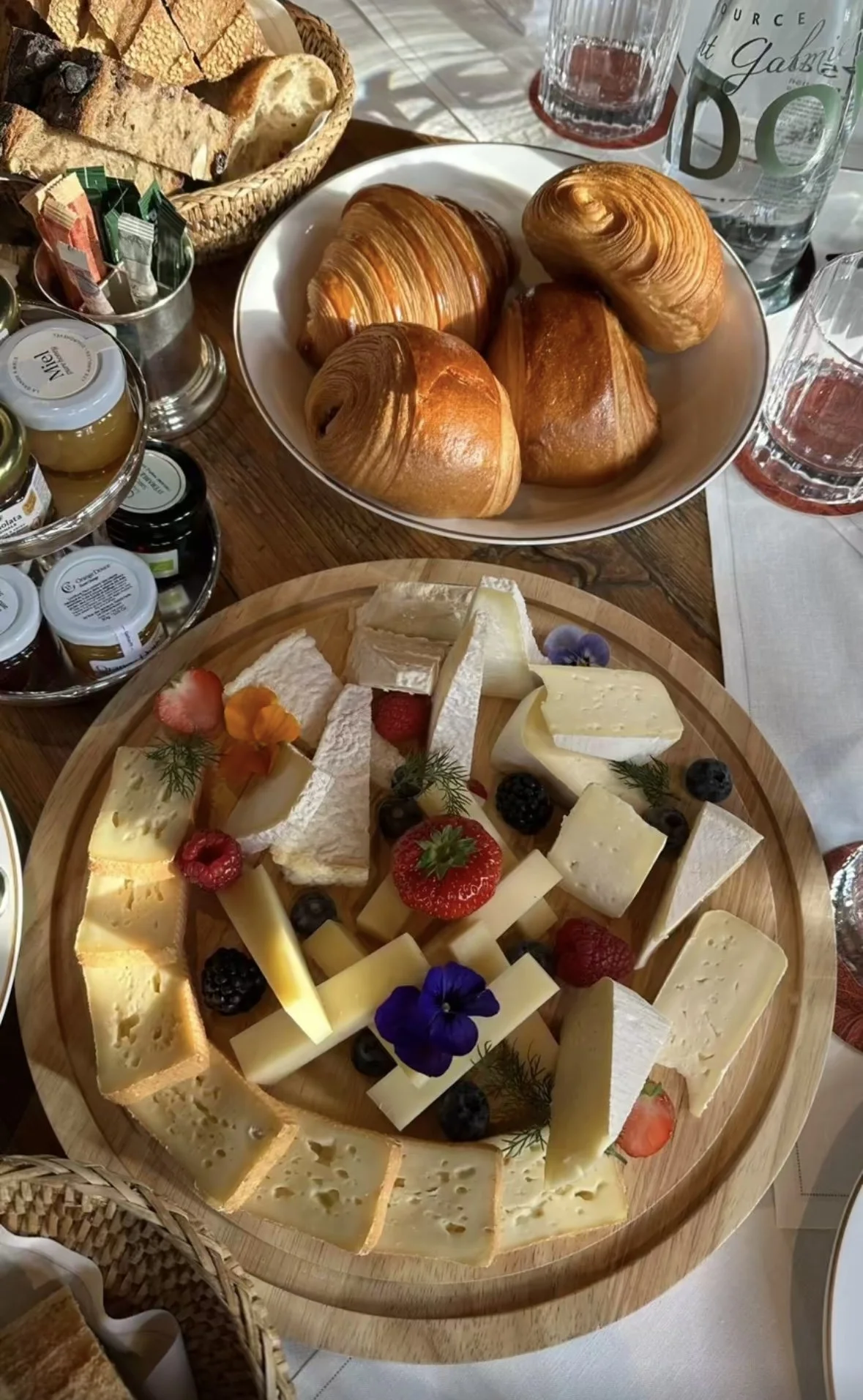 Plate de fromages variés avec fruits frais, croissants et pains dans une assiette, avec des verres d'eau et des pots de confitures sur la table.