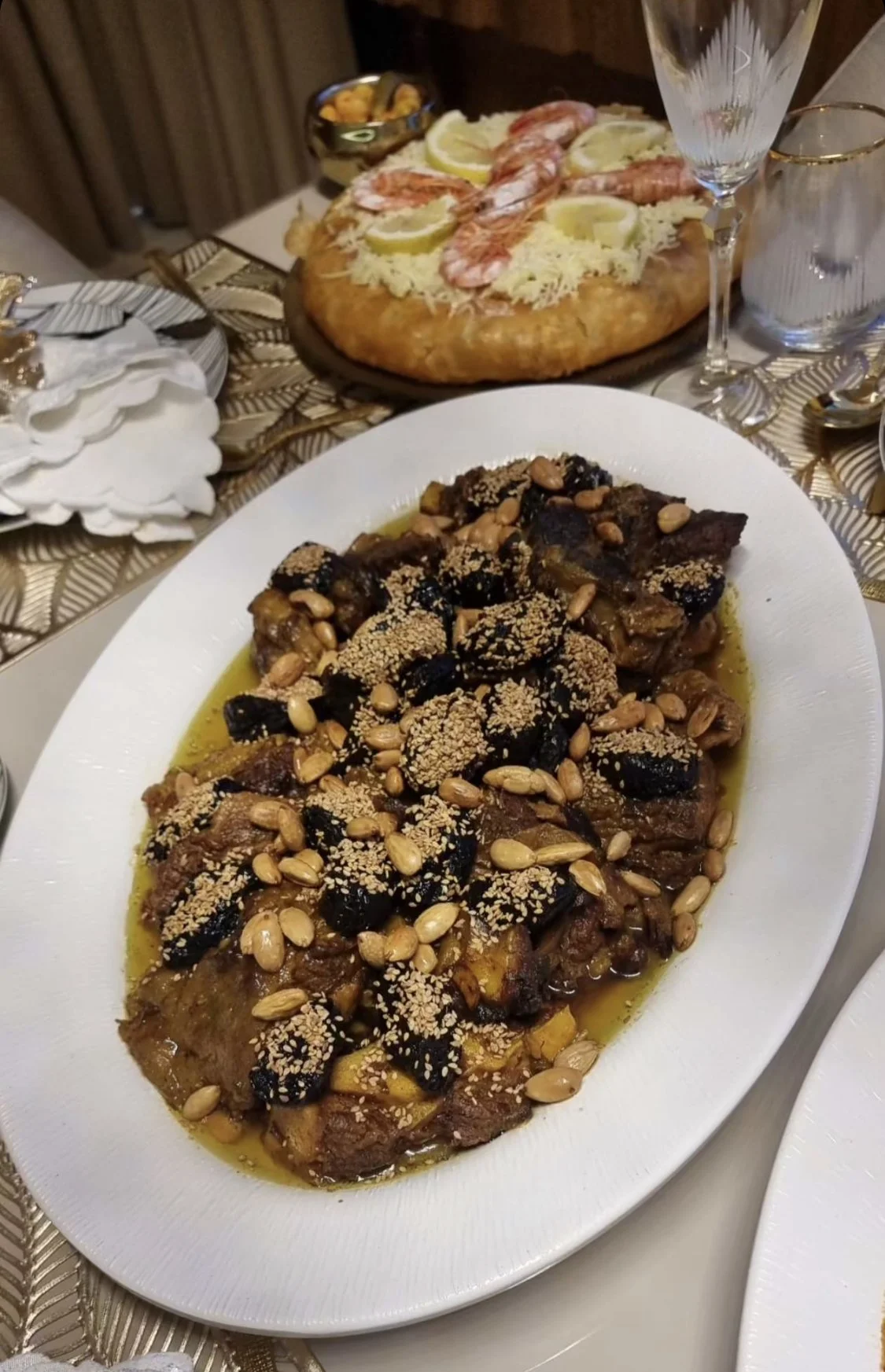 Plat principal de viande en sauce garnie de graines et légumes, avec une pizza en arriêre plan, accompagnée de vin, verre et assiettes, sur une table avec nappe.