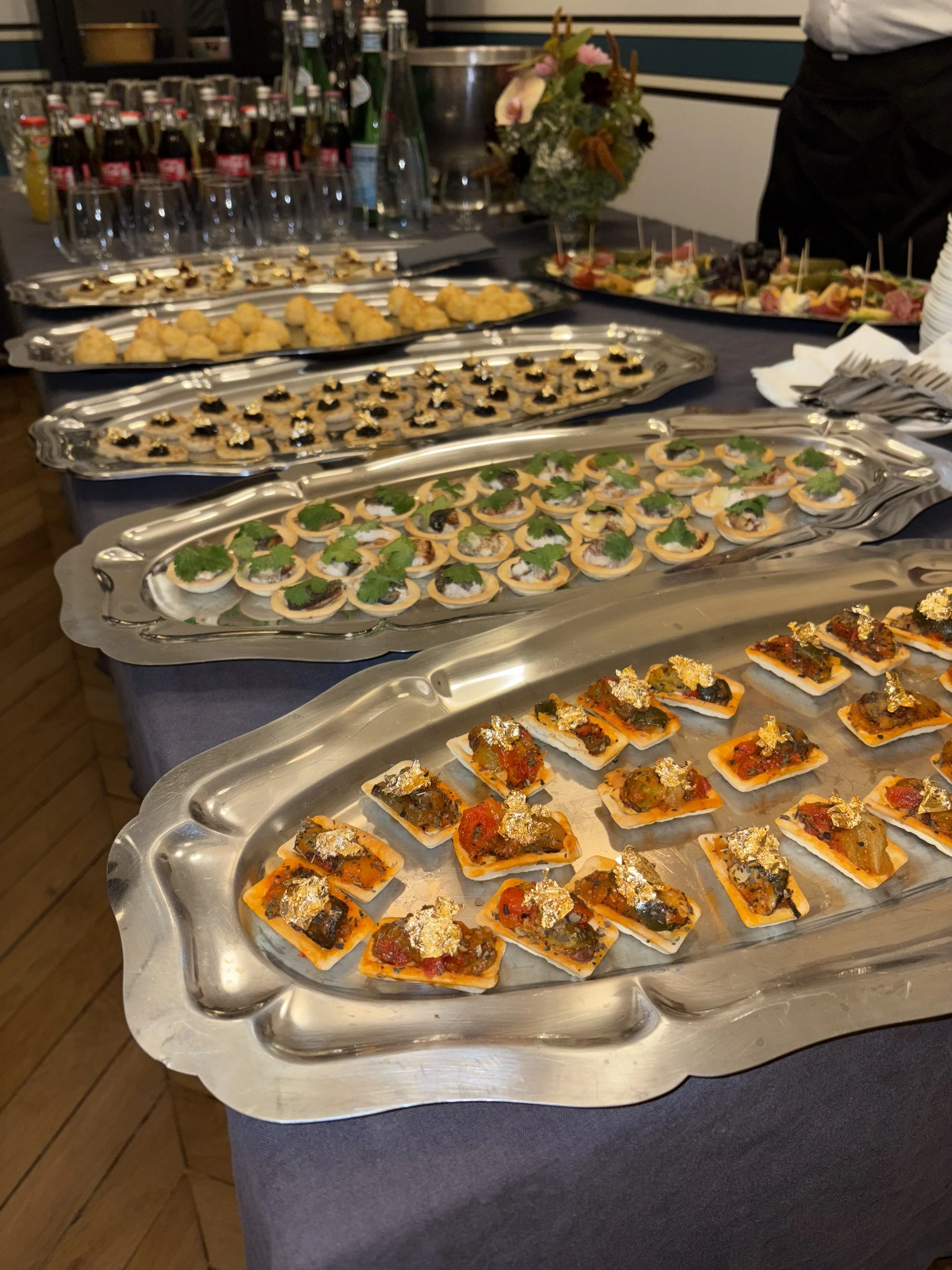 Plateaux de canapés variés lors d'un buffet, avec des cuillères et des fleurs en arrière-plan.