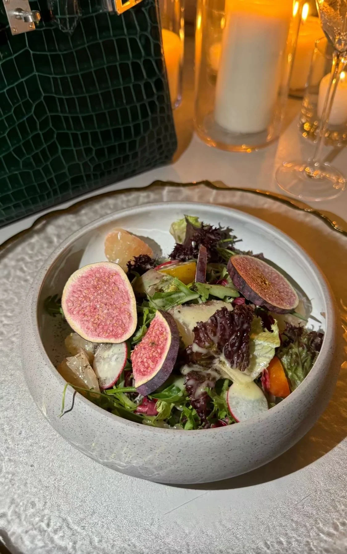 Salade composée avec des tranches de figues, des feuilles de salade et d'autres légumes dans un bol blanc, placé sur une table avec des bougies et une sacoche à l'arrière.