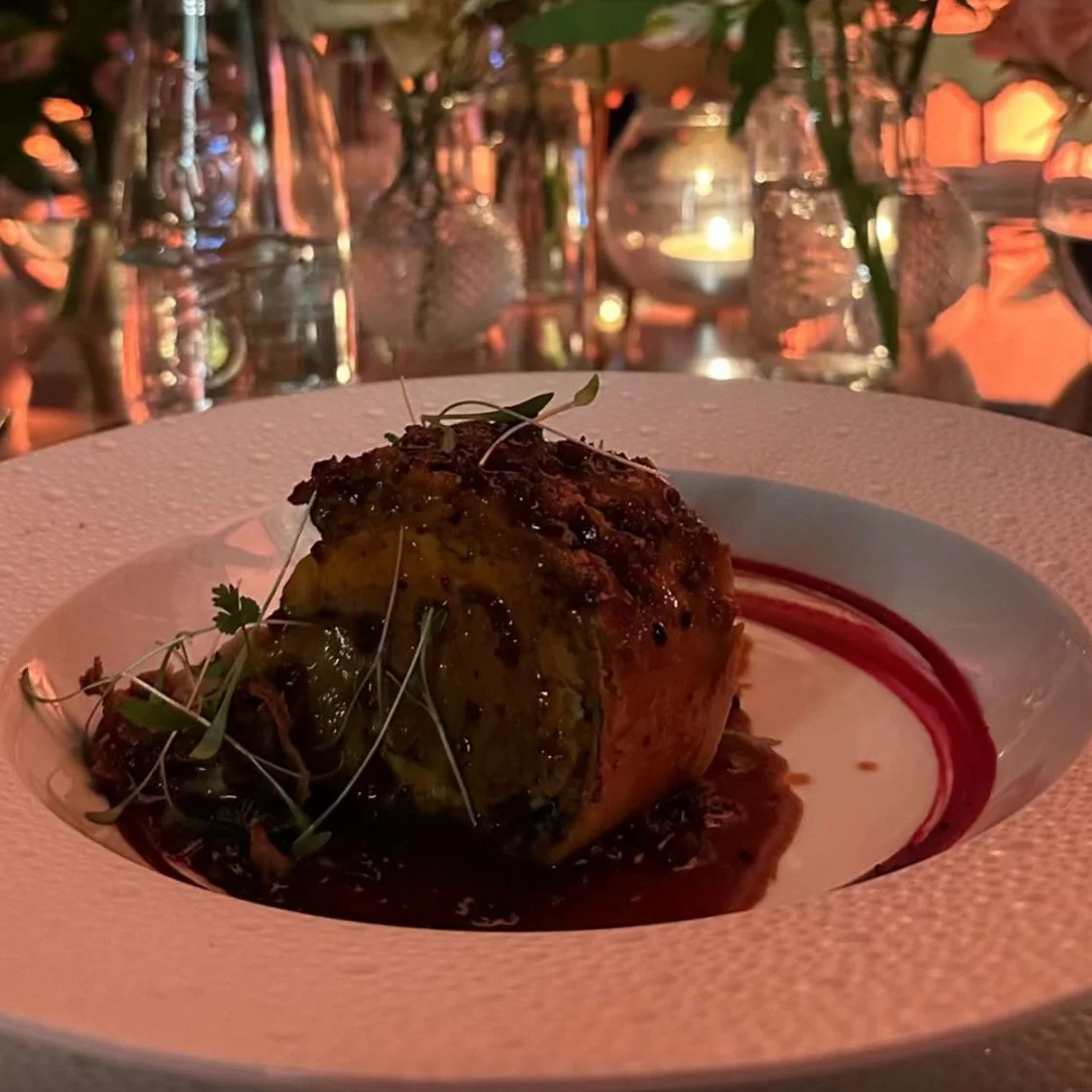 Plat de viande ou de poisson avec sauce, décoré de microgreens, dans un restaurant élégant avec des bougies et des verres en arrière-plan.