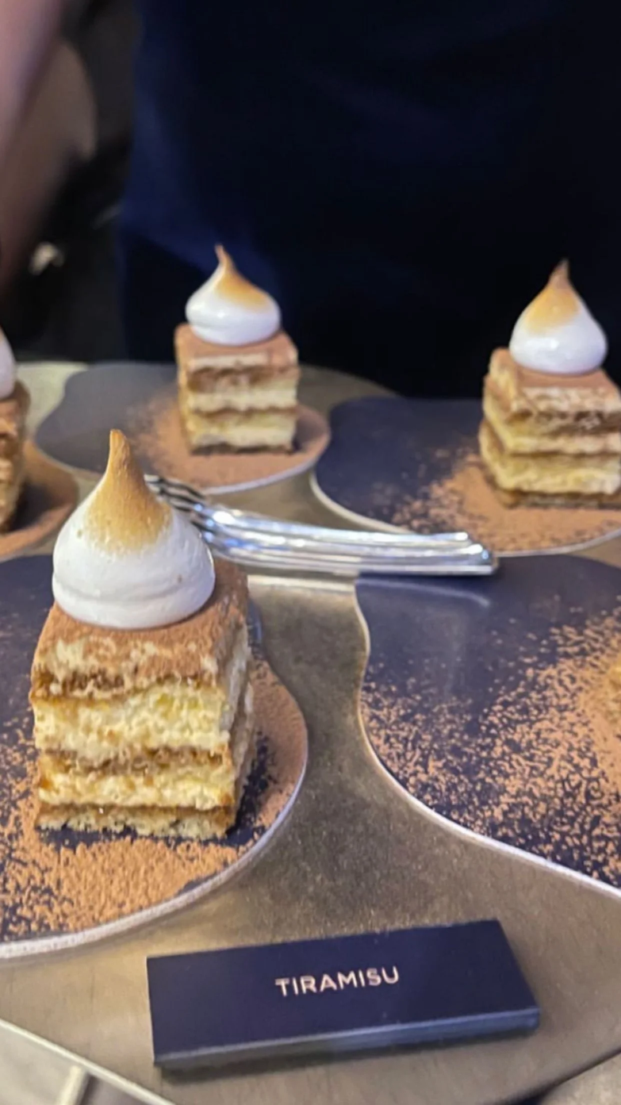 Trois parts de gâteau Tiramisu avec mousse en forme de pointe dorée, posées sur des assiettes en laiton avec une poudre de cacao autour, dans un buffet de dessert.