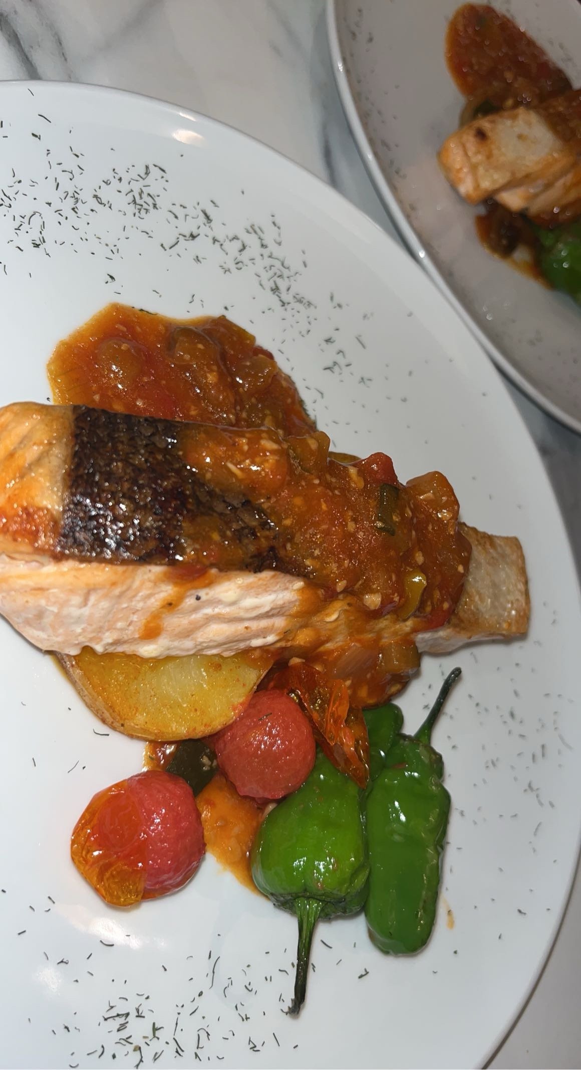 Pièce de poisson grillé servie avec une sauce tomate, accompagné de pommes de terre, de tomates cerises, et de piments verts.