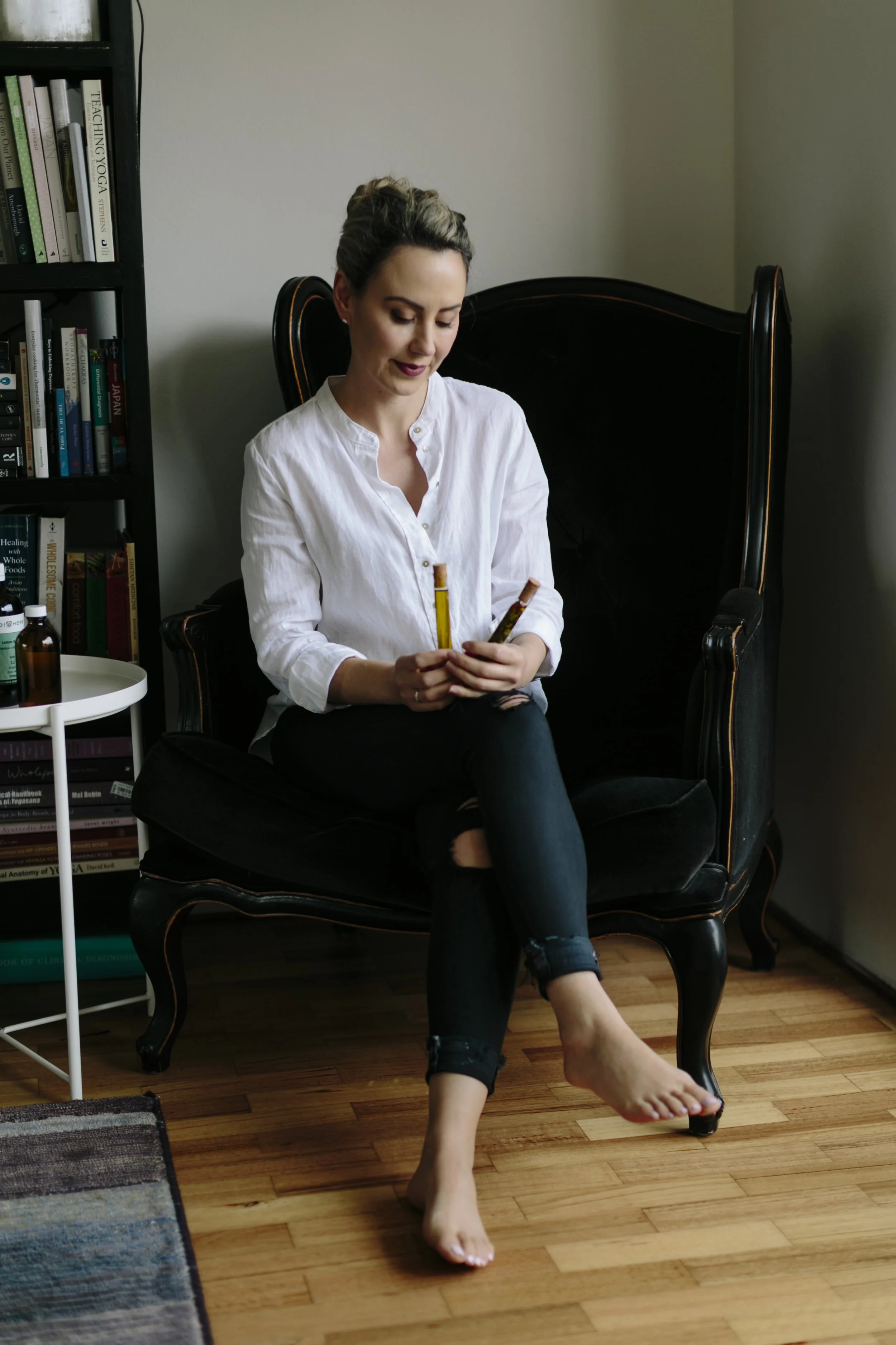 A woman sitting on a black vintage armchair with a high back, holding a paintbrush and a yellow pencil, in a room with a bookshelf, wooden flooring, and a white side table with a bottle. The woman is wearing a white shirt and black pants, with one leg crossed over the other, barefoot, with her hair styled up and a contemplative expression.