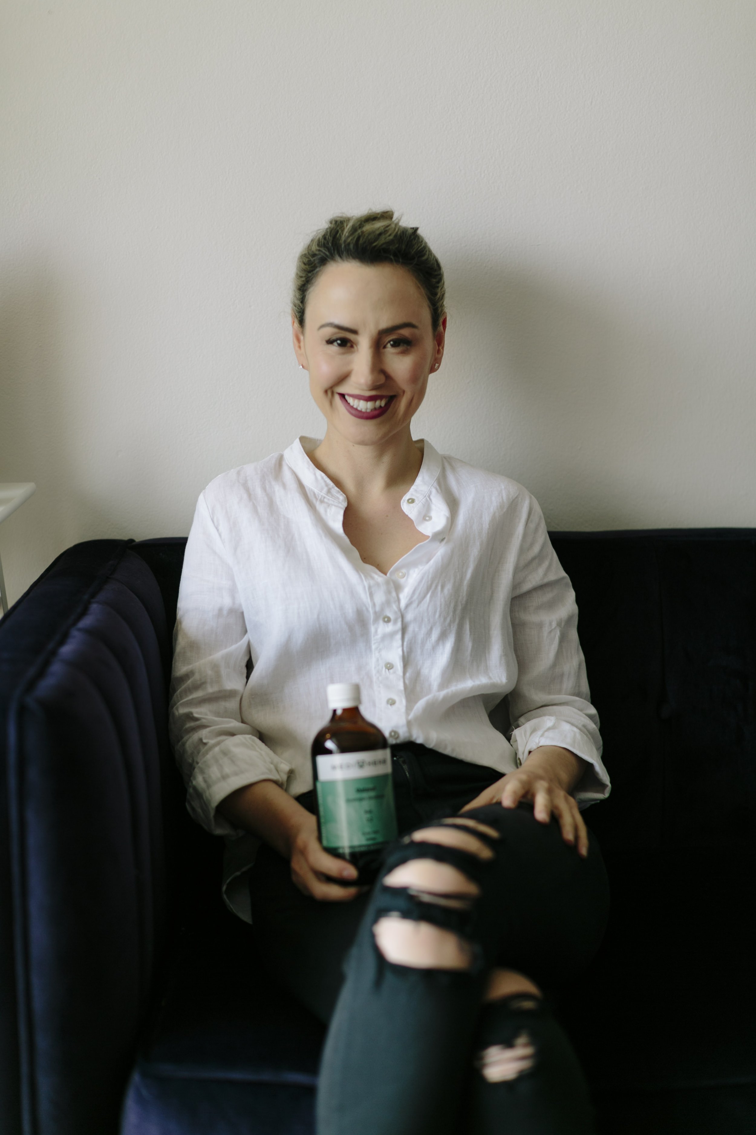 A smiling woman with dark hair tied back, wearing a white button-up shirt, black ripped jeans, and black sandals, sitting on a black couch against a plain light-colored wall. She is holding a medicine bottle in her right hand.