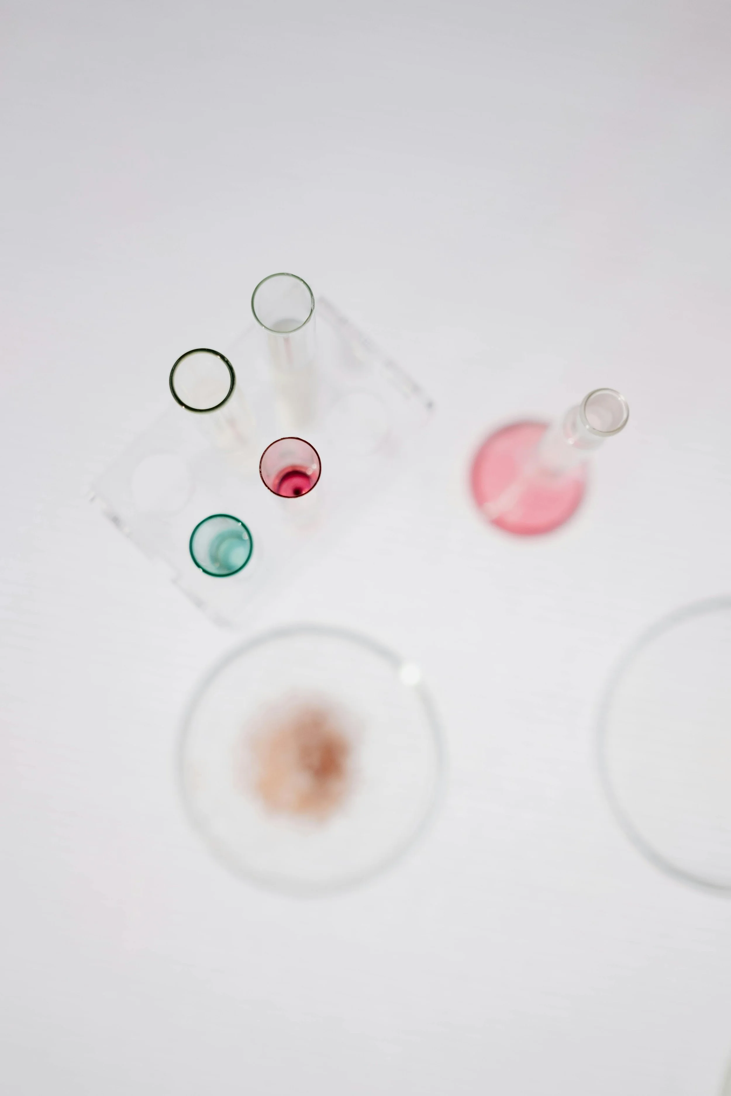 Multiple test tubes with colored liquids on a white surface, some with pink, green, and clear liquids, with shadows cast on the surface.