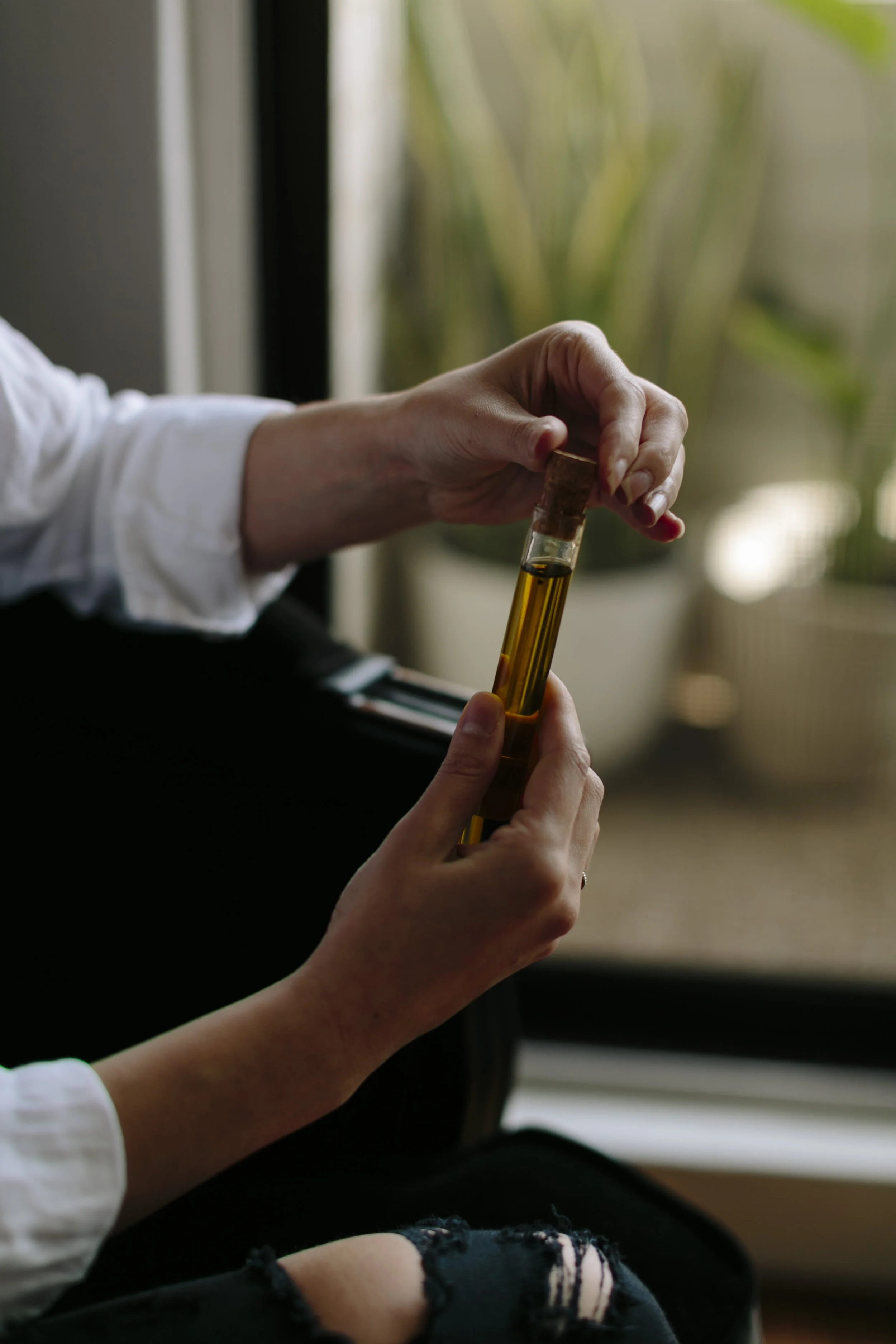 Close-up of a person's hands holding a small glass container with a cork lid, filled with a yellowish liquid, sitting near a window with blurred green plants in the background.