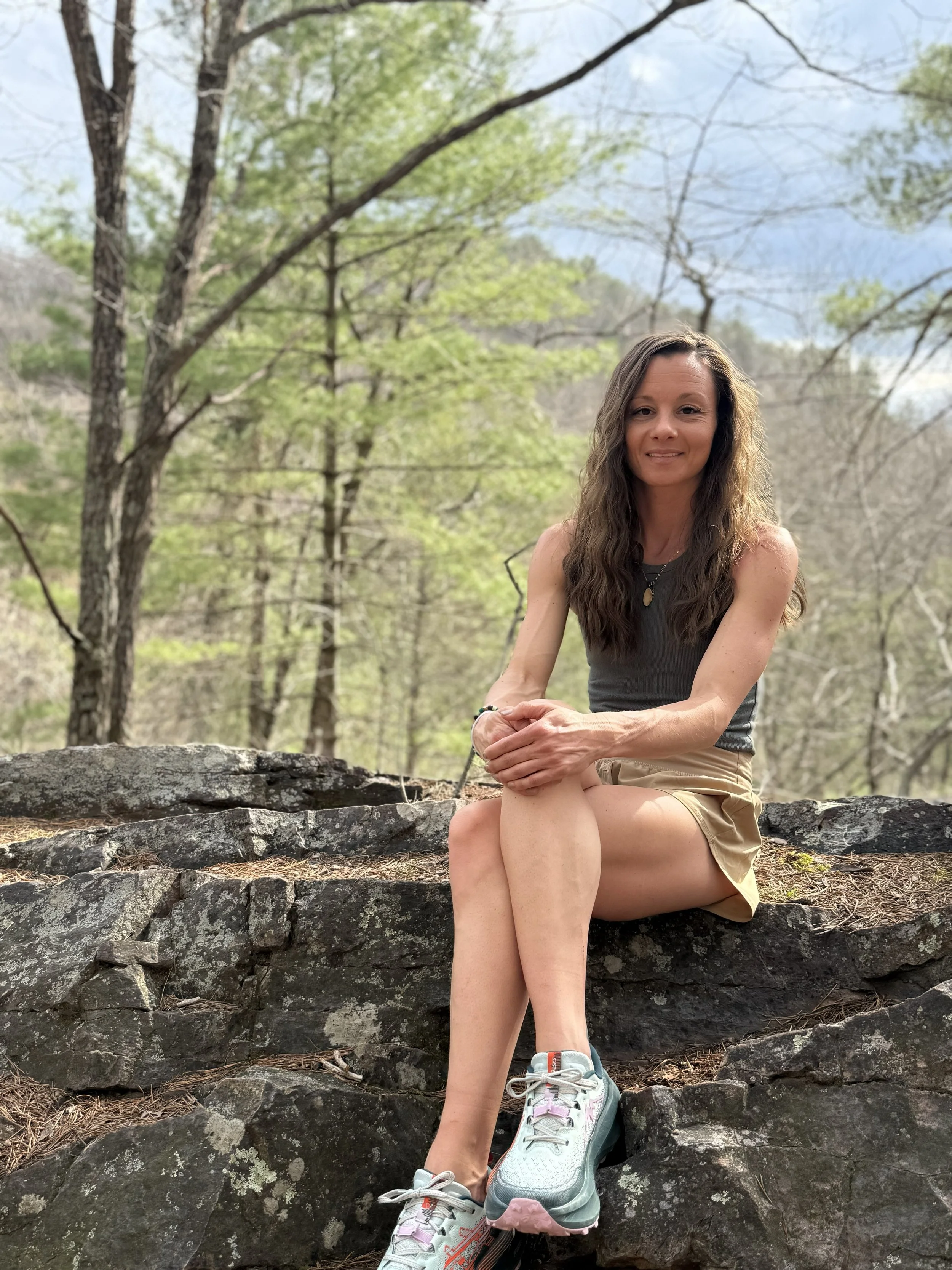 A woman with long wavy hair sitting on a large rock outdoors, surrounded by trees with budding leaves, wearing a gray sleeveless top, beige shorts, and hiking shoes, smiling at the camera.