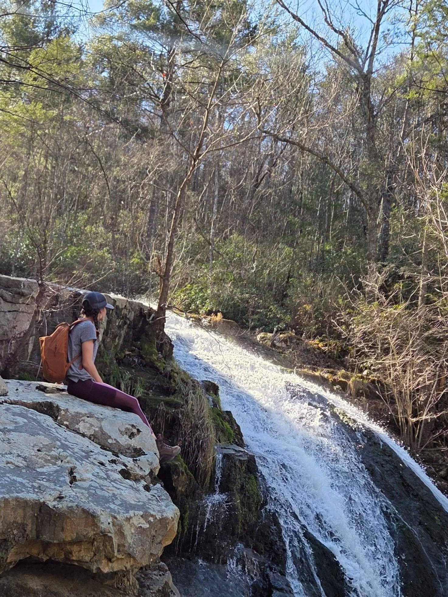 A person sitting on rocks beside a waterfall in a wooded area, wearing a cap, backpack, and outdoor clothing.