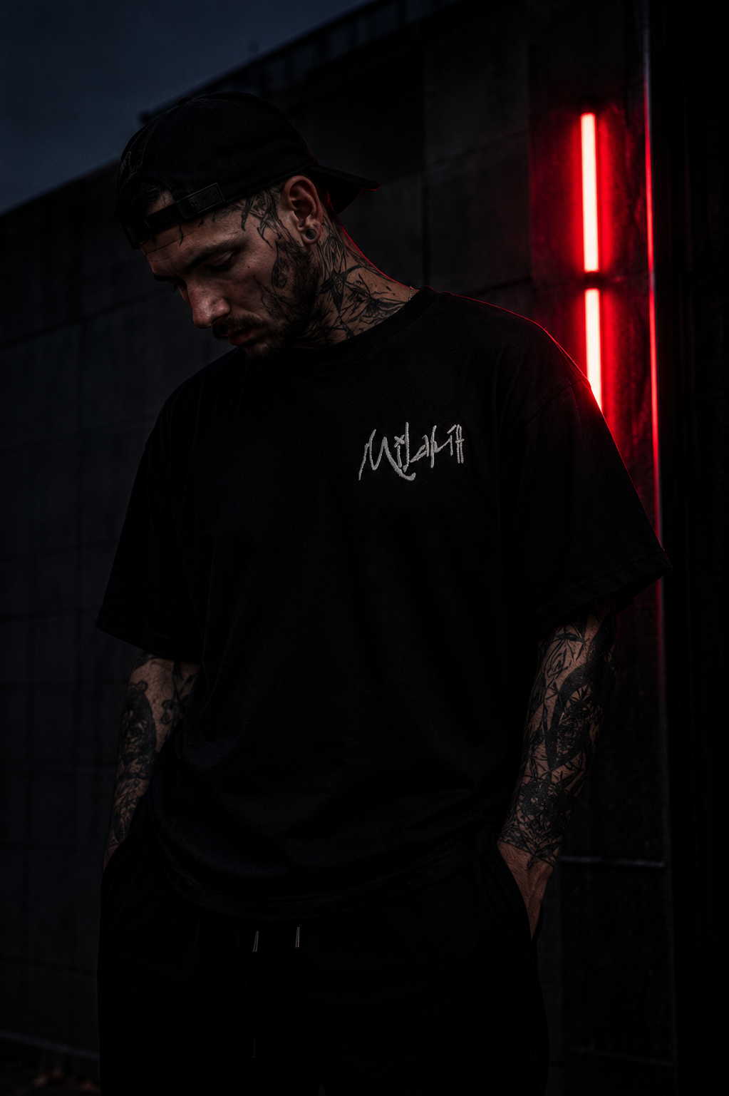 Junger Mann mit Tattoos, schwarzer Kleidung und schwarzer Kappe, steht bei Nacht vor einer schwarzen Wand mit rotem Neonlicht