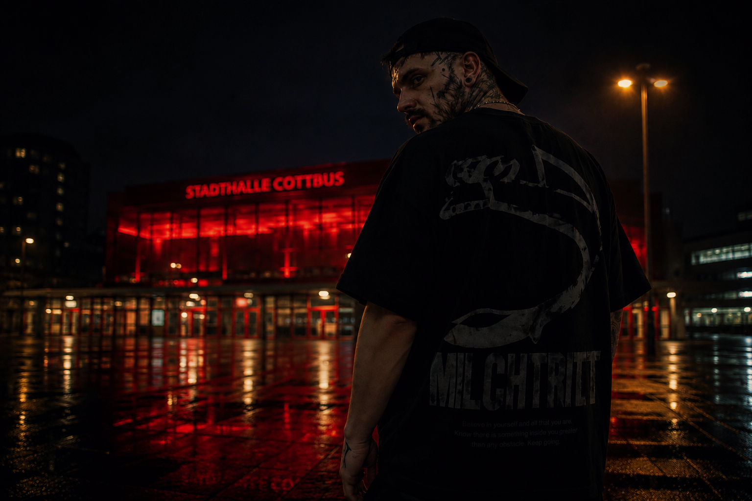 Junger Mann mit Tattoos bei Nacht vor der Stadthalle Cottbus, beleuchtet von roten Lichtern, nasse Straße reflektiert das Licht.