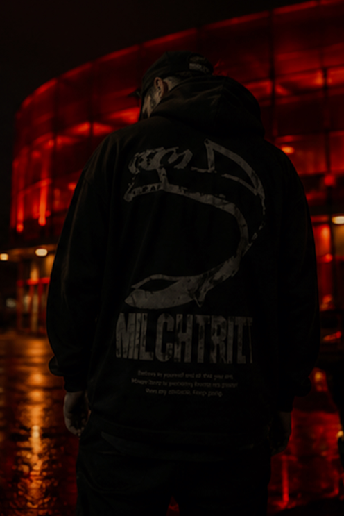 Junge Person trägt dunkle Jacke mit Graffiti-Design, Hintergrund rot leuchtendes Gebäude bei Nacht.